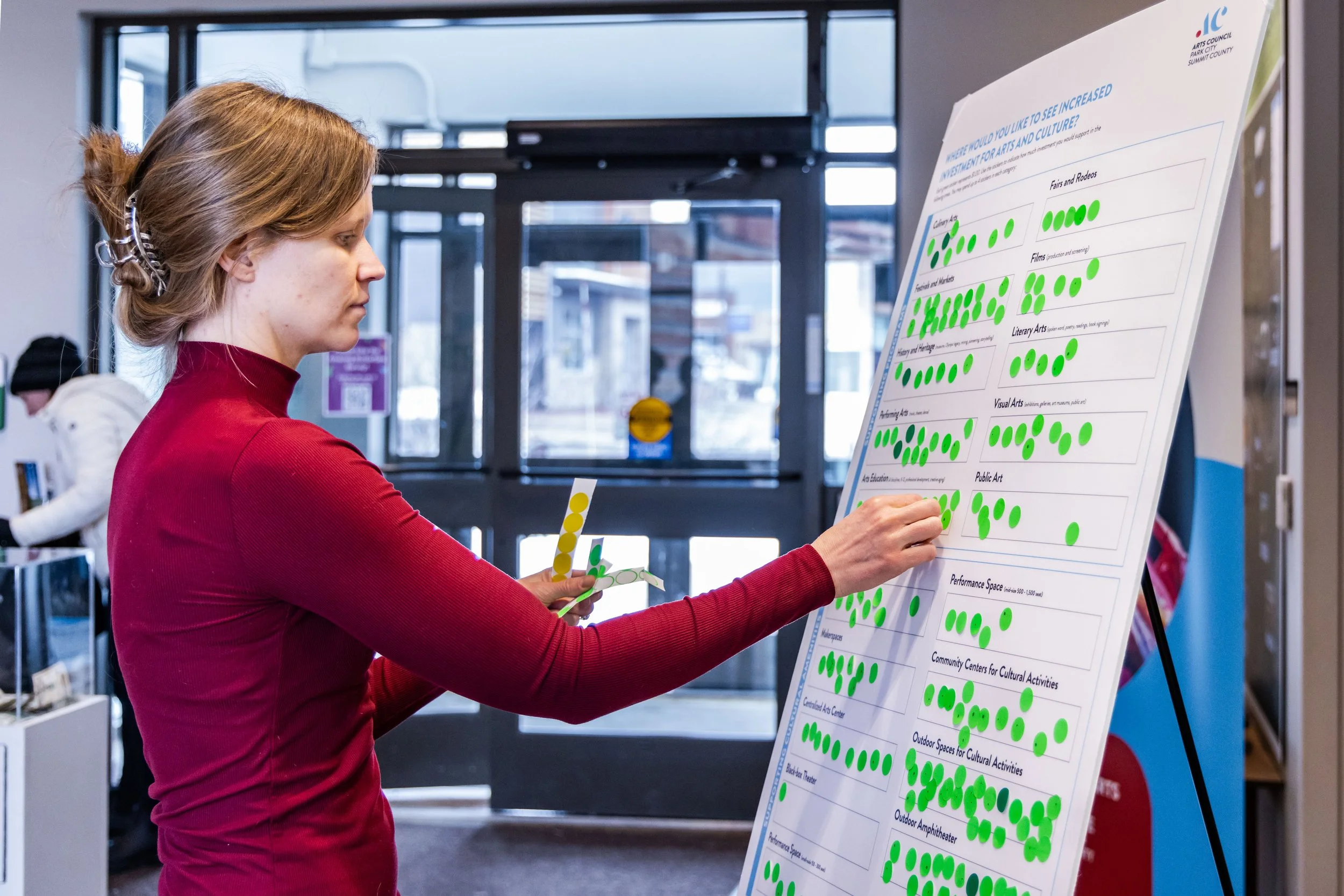 A woman in a red long-sleeve shirt pins green dots on a large survey board about community interests and cultural spaces inside a building with glass doors.