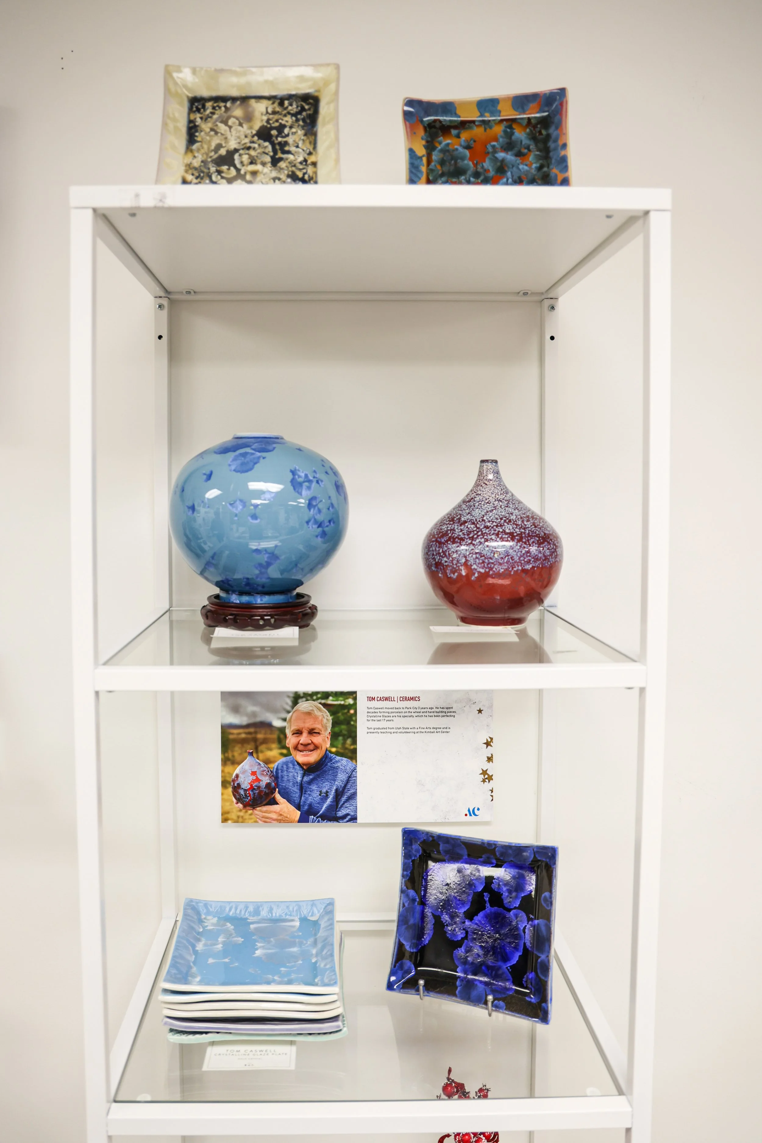 Display case with blue and red ceramic vases and plates, including a blue round vase, a red and white speckled vase, and patterned plates on top, with a photo of a man holding one of the vases.