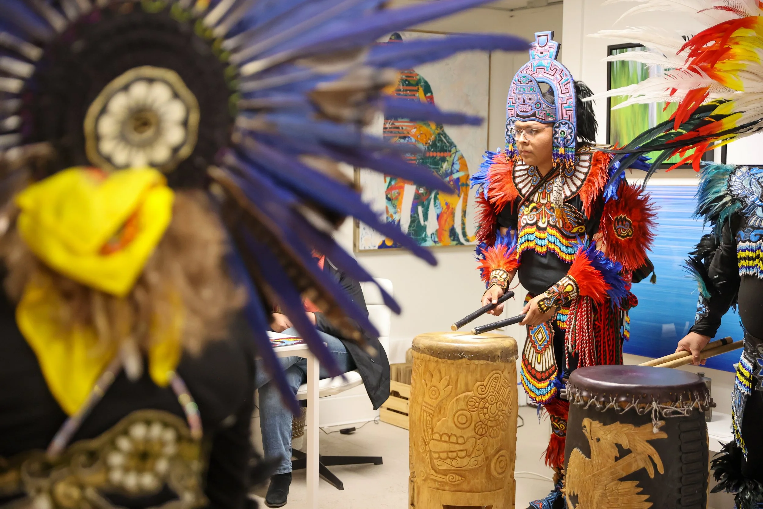 Uma pessoa vestida com trajes indígenas tradicionais, incluindo um cocar colorido com penas e acessórios com miçangas, toca tambores cerimoniais num ambiente interno. Outras pessoas com trajes semelhantes são parcialmente visíveis, e obras de arte estão expostas. 