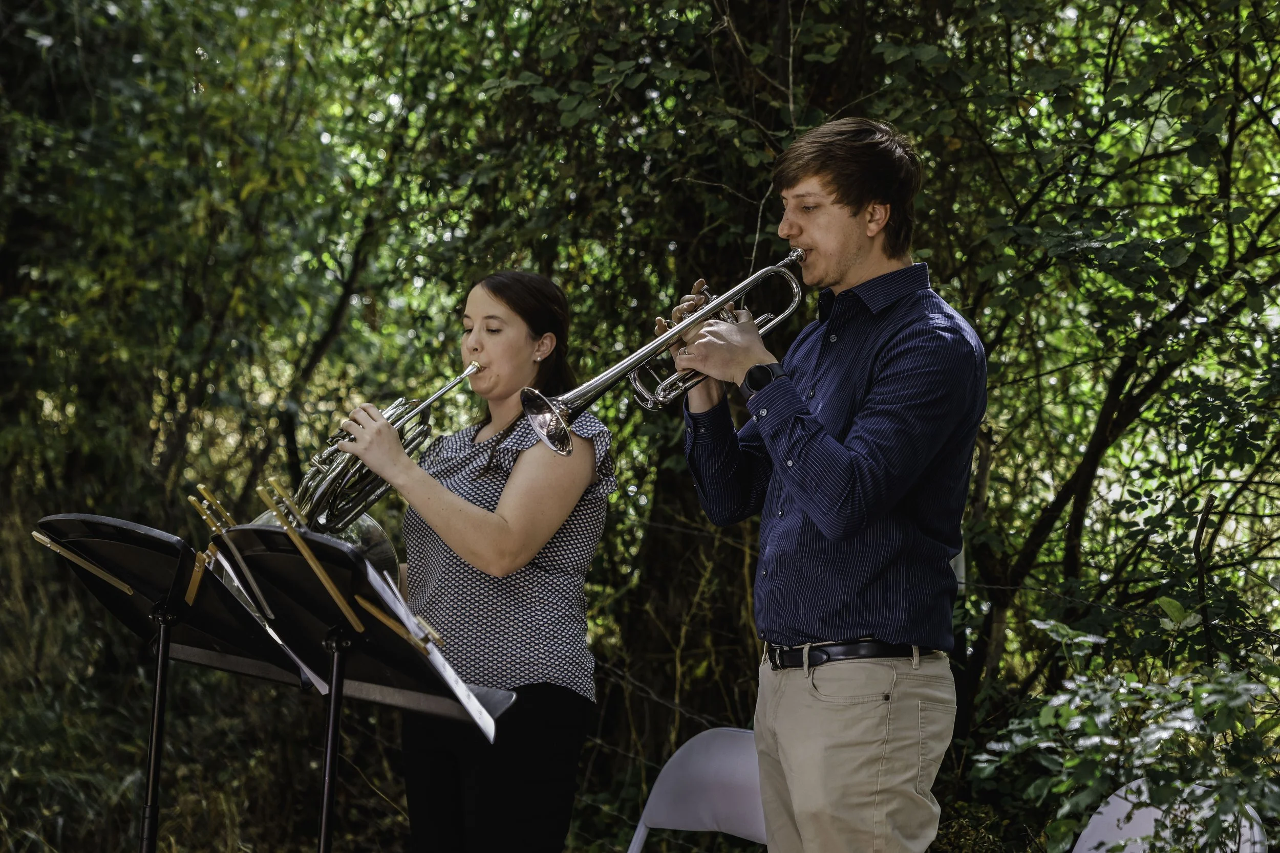 Uma mulher e um homem a tocar clarinete e trompete ao ar livre, entre árvores