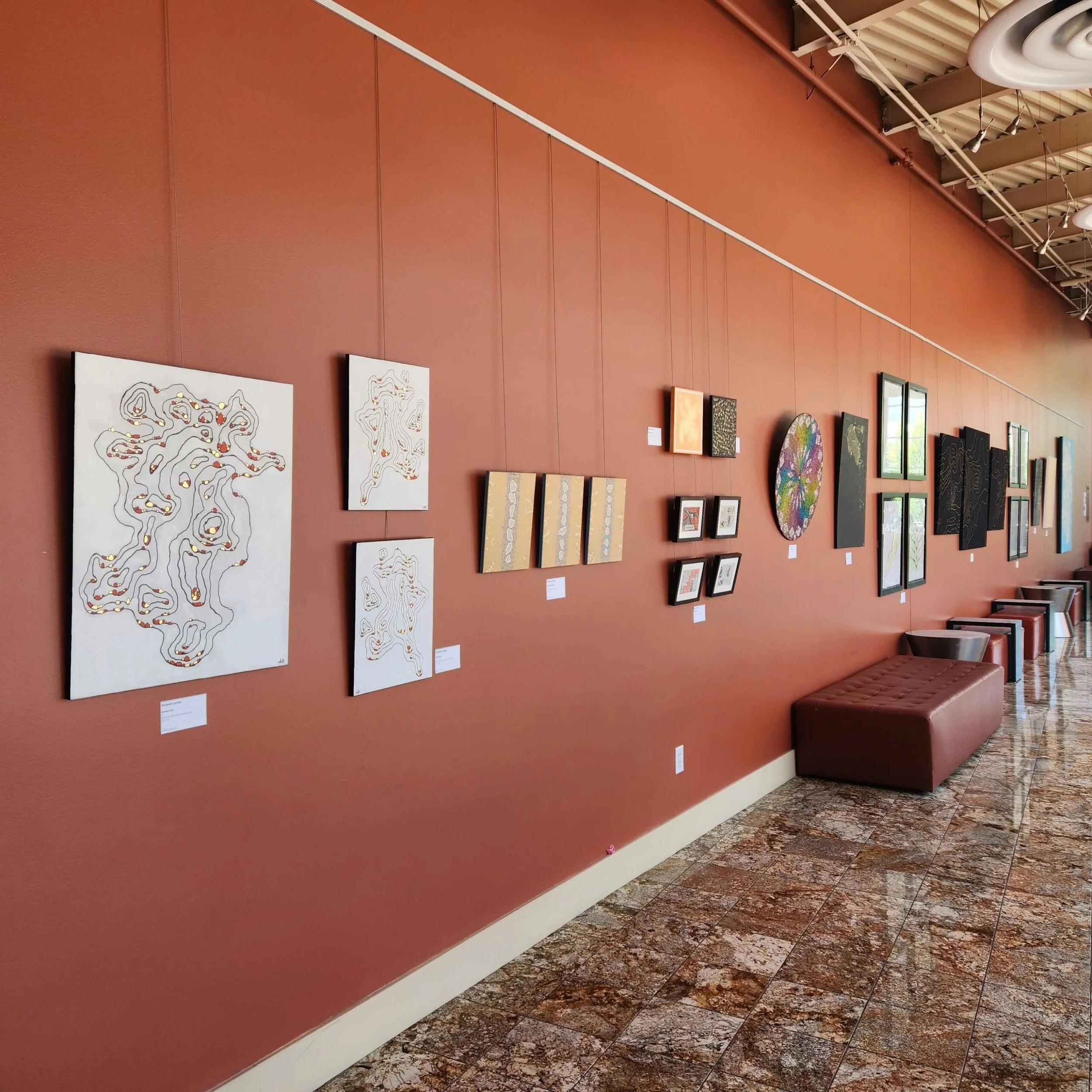 Uma galeria de arte com uma parede cor de ferrugem exibindo várias pinturas abstratas e obras de arte emolduradas, com assentos ao longo da parede e um piso de mármore polido.