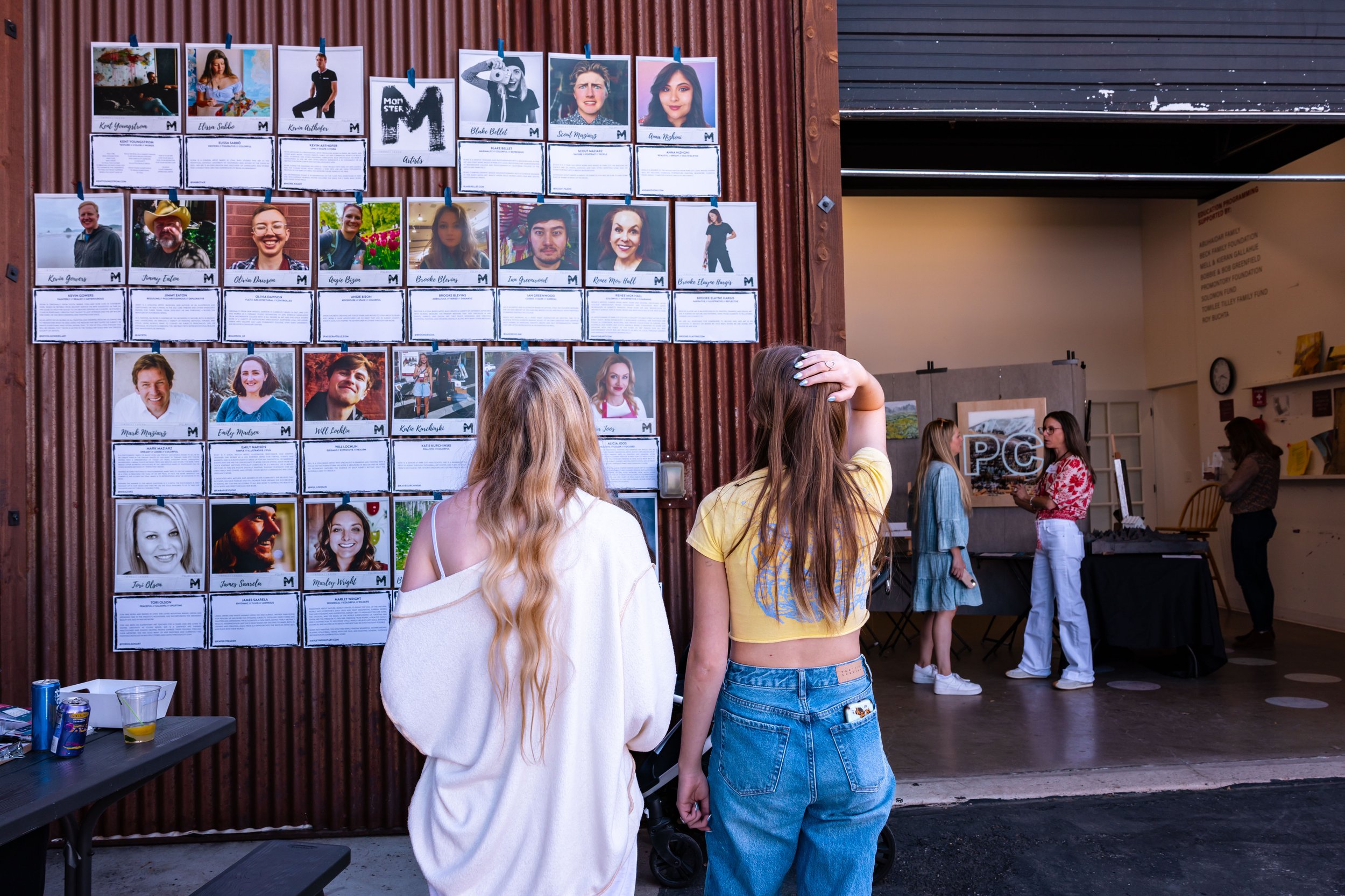 People observing a wall with photographs and descriptions at an indoor event or exhibit, with additional individuals interacting at a table in the background.
