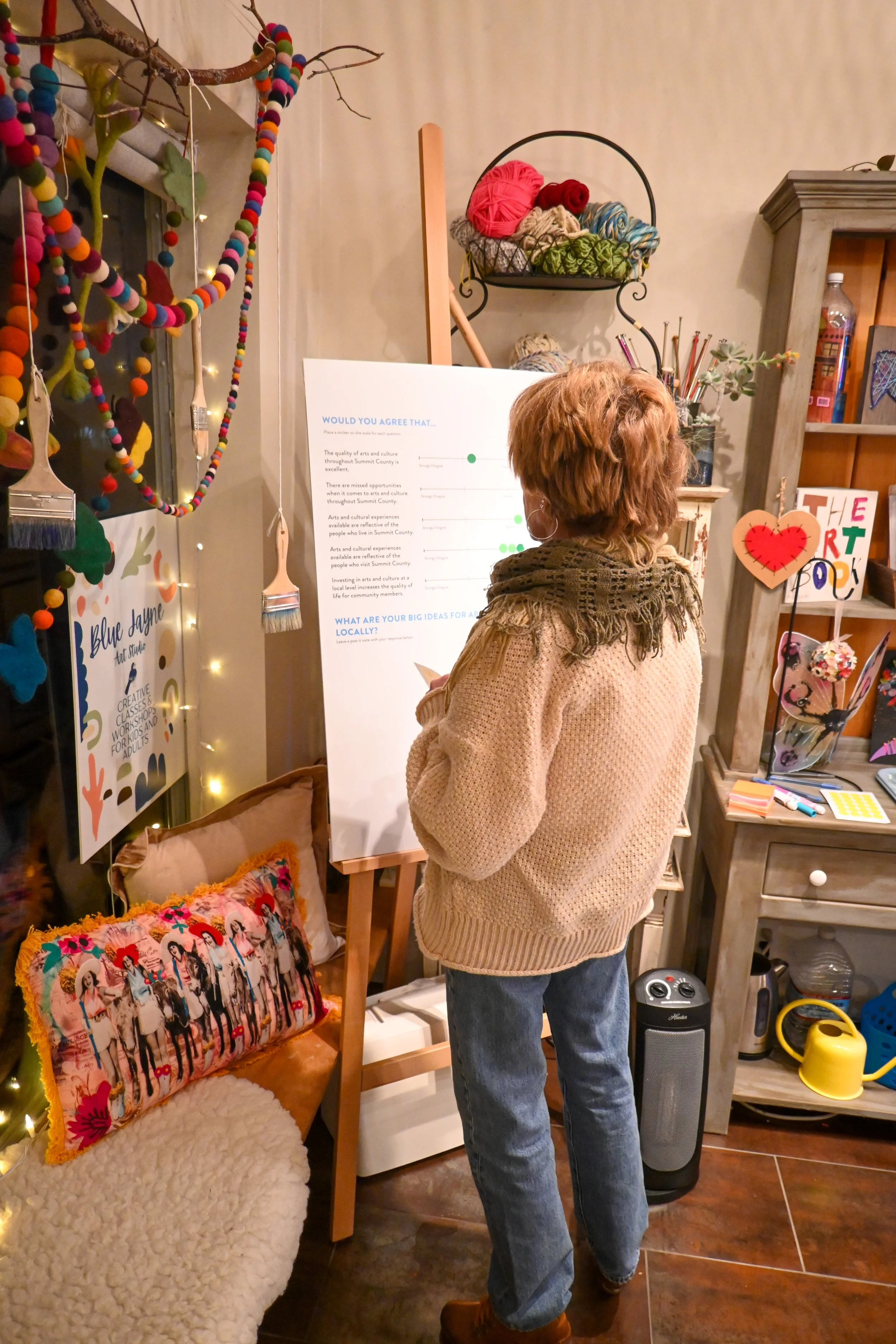 Woman with curly hair and glasses reading a poster at a decorated indoor event, with colorful artwork, yarn, and crafts around her.