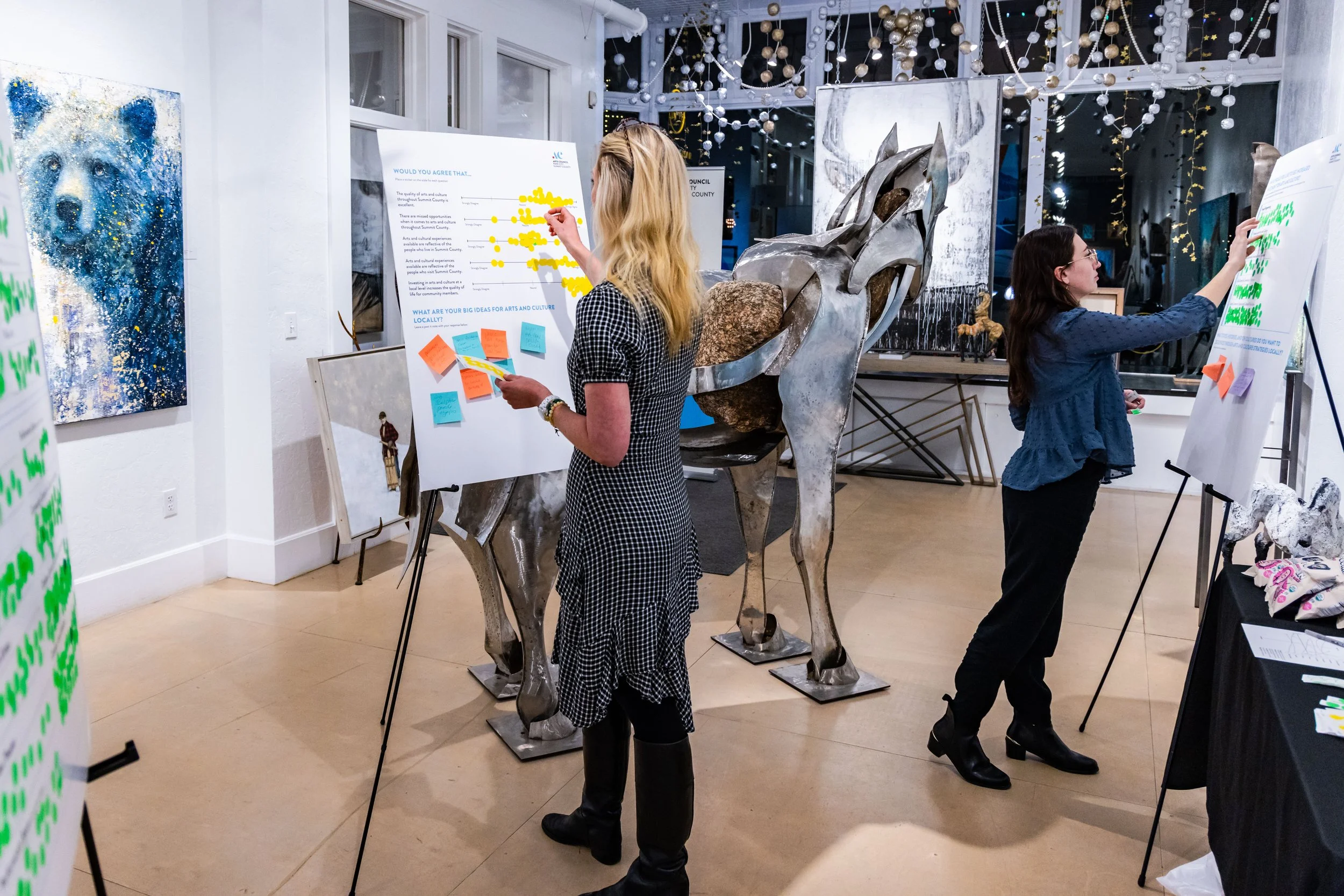 Two women standing at presentation boards, engaged in a voting activity, in an art gallery with hanging decorations, artwork, and a metallic horse sculpture in the background.