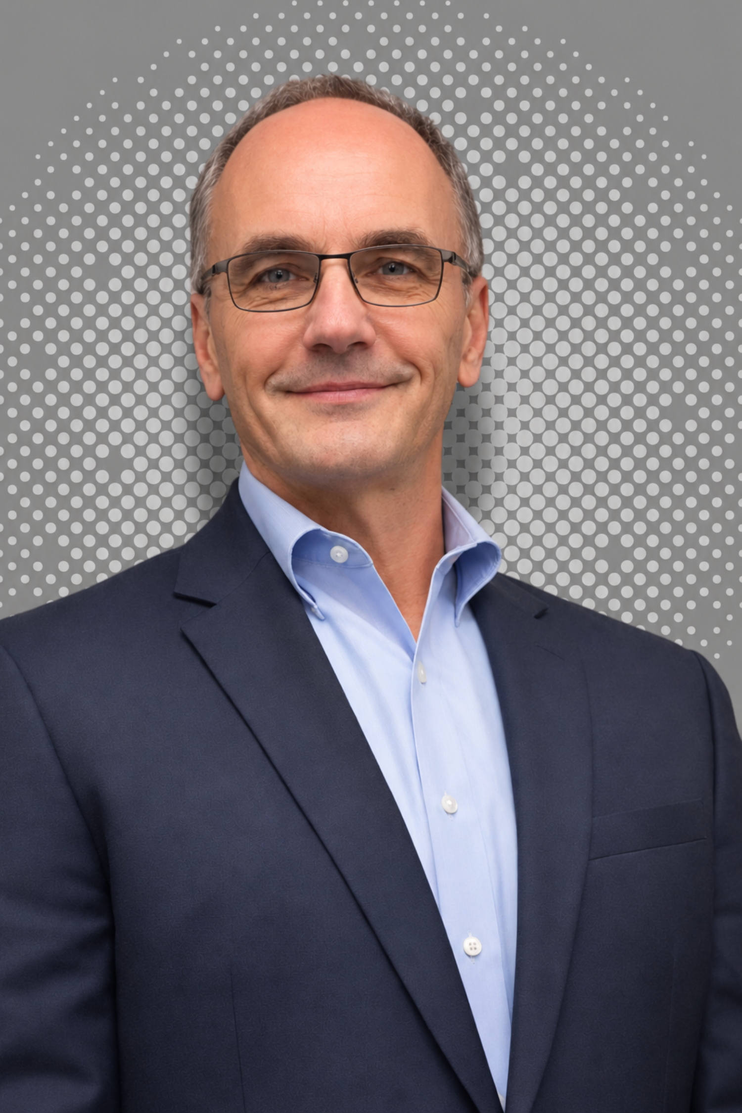 A middle-aged man with glasses, dressed in a dark suit and light blue shirt, smiling in front of a gray background with white dots.
