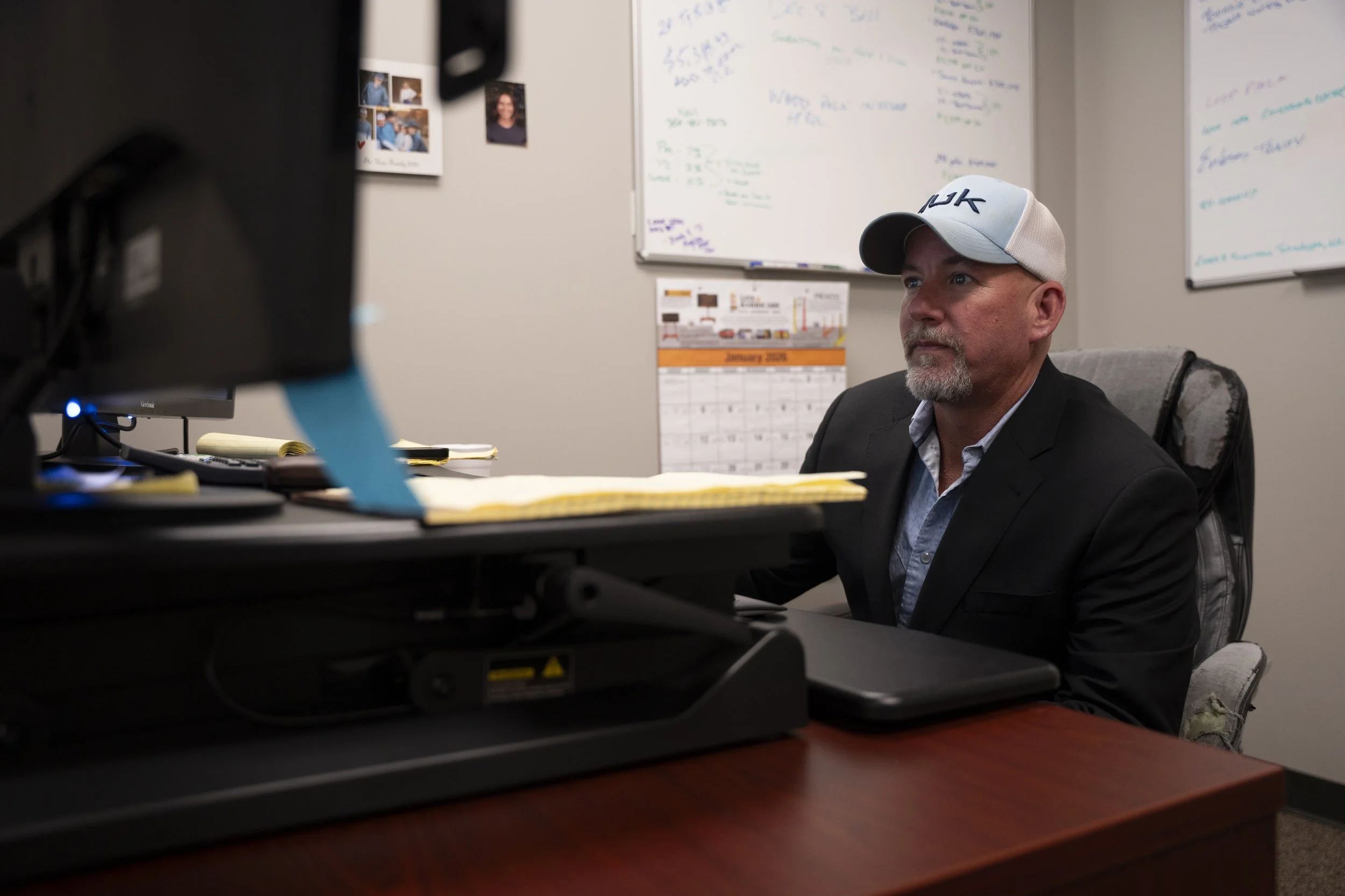 A man with a beard and mustache wearing a gray baseball cap, a dark blazer, and a light shirt, sitting at a desk in an office. Behind him, there is a whiteboard with handwritten notes, a calendar, and some photographs on the wall.