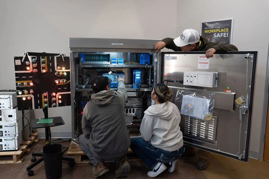 Three technicians working on an open electrical control panel in an industrial or educational setting, with a safety poster on the wall.