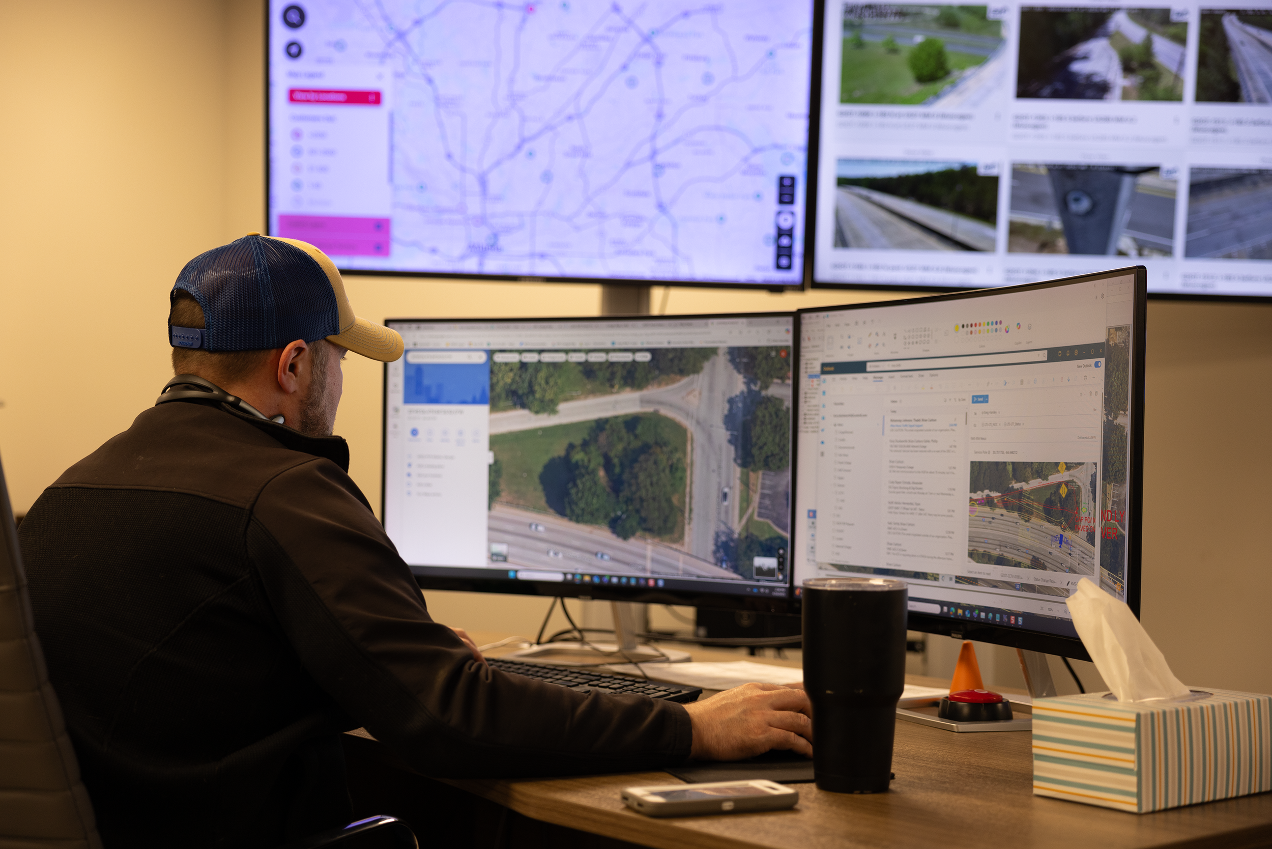 A person wearing a blue and beige cap working at a desk with multiple computer monitors displaying maps, traffic data, and photography of roads and highways, in an office setting.