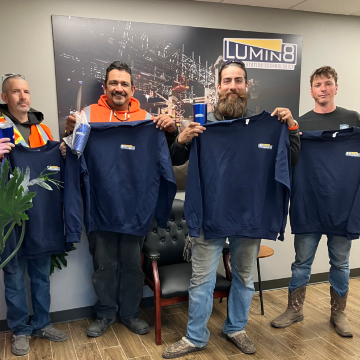 Four men standing indoors, holding navy blue shirts with the Lumin8 logo, in front of an office wall.