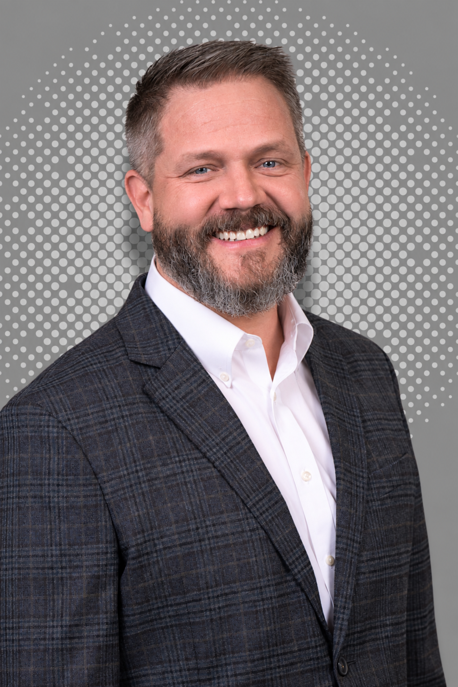 Headshot of a smiling man with short, dark hair, a beard, and wearing a checkered dark blazer and white shirt, against a dotted gray background.