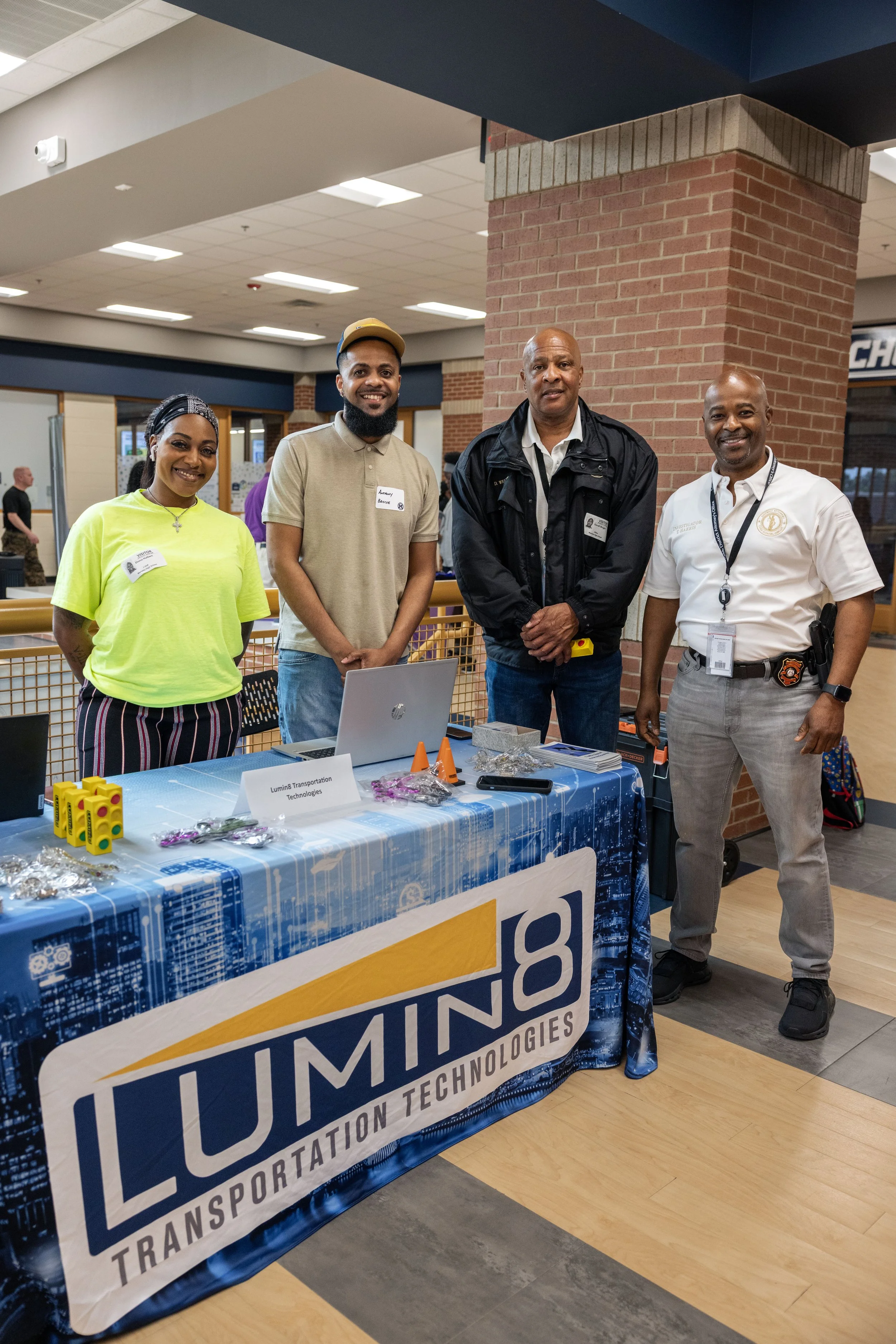 Four people standing behind a table at an indoor event, with a display sign reading 'Lumin8 Transportation Technologies'. The table has various items, including yellow devices, purple objects, a laptop, and informational materials. The people are smi