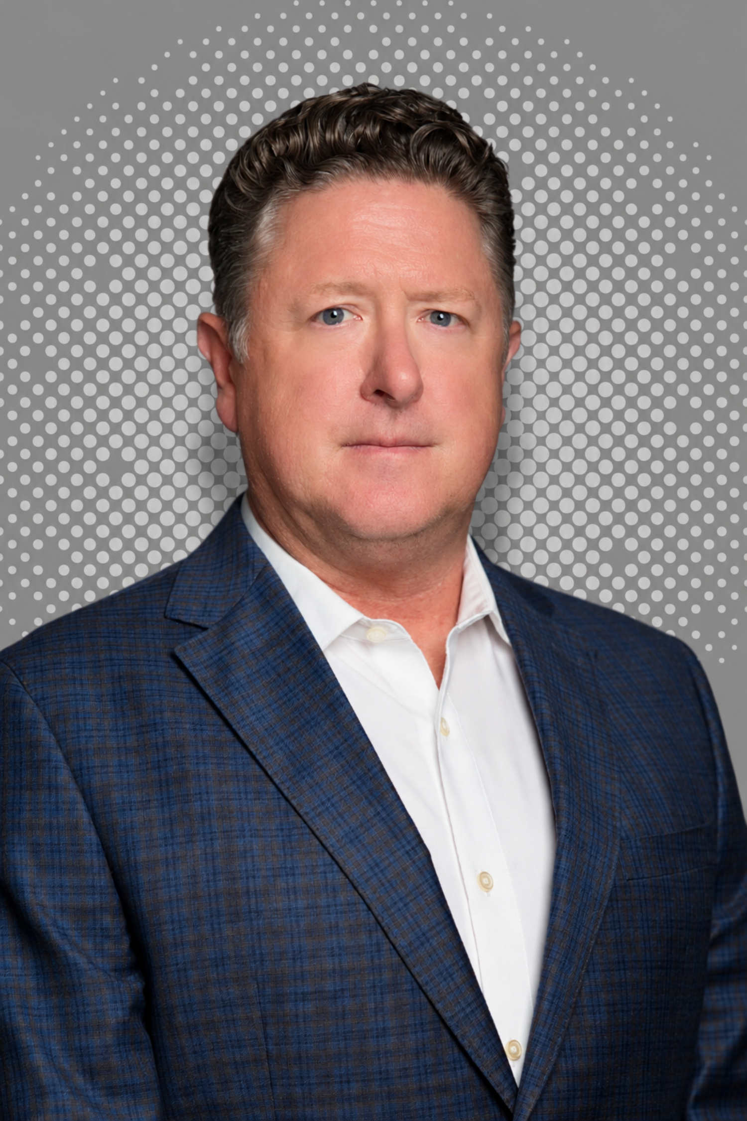 A middle-aged man with blue eyes and short, dark, wavy hair wearing a blue checked suit and white shirt, standing against a gray background with white dots.