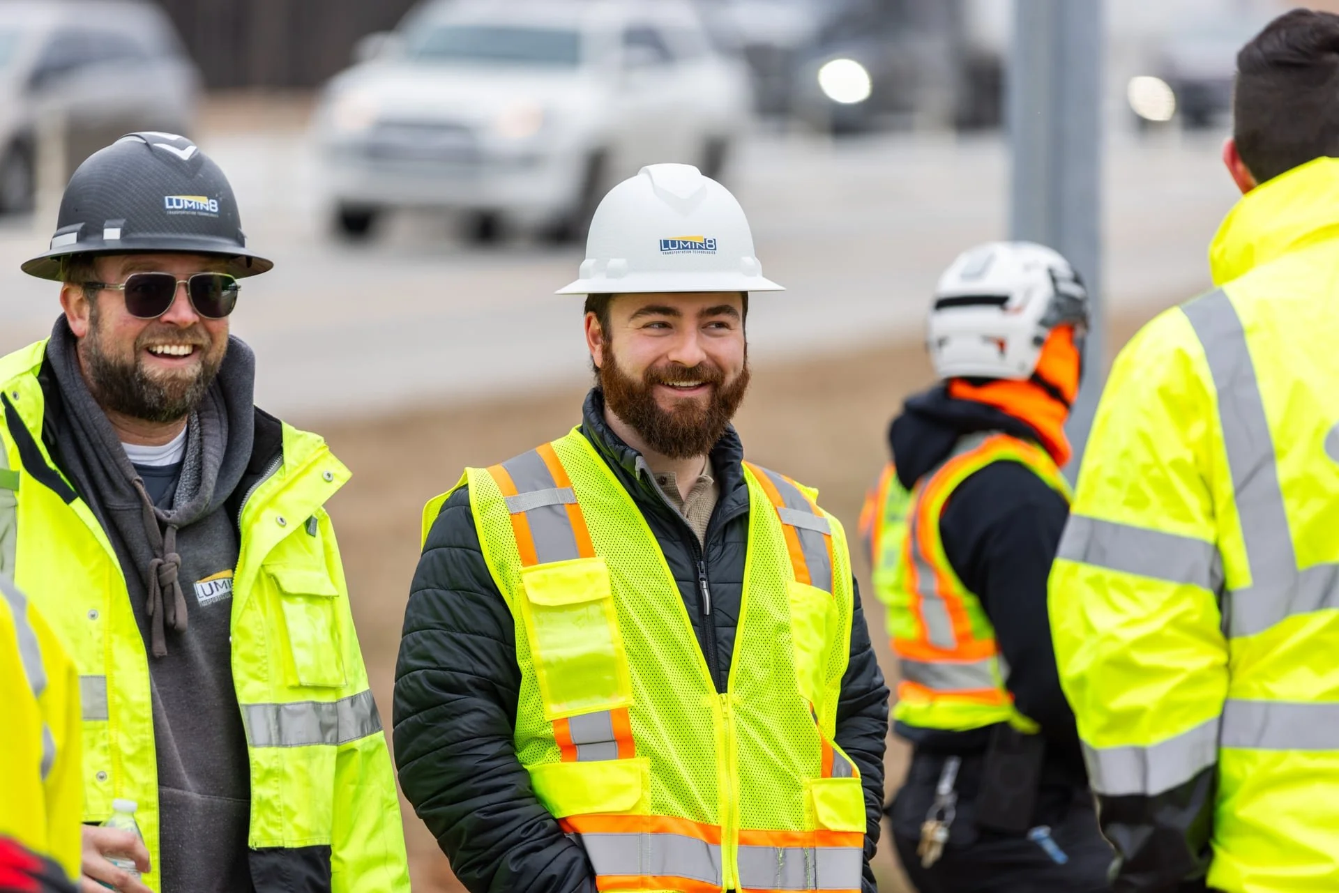 Group of construction workers wearing high-visibility safety vests and hard hats standing outdoors, with blurred cars in the background.