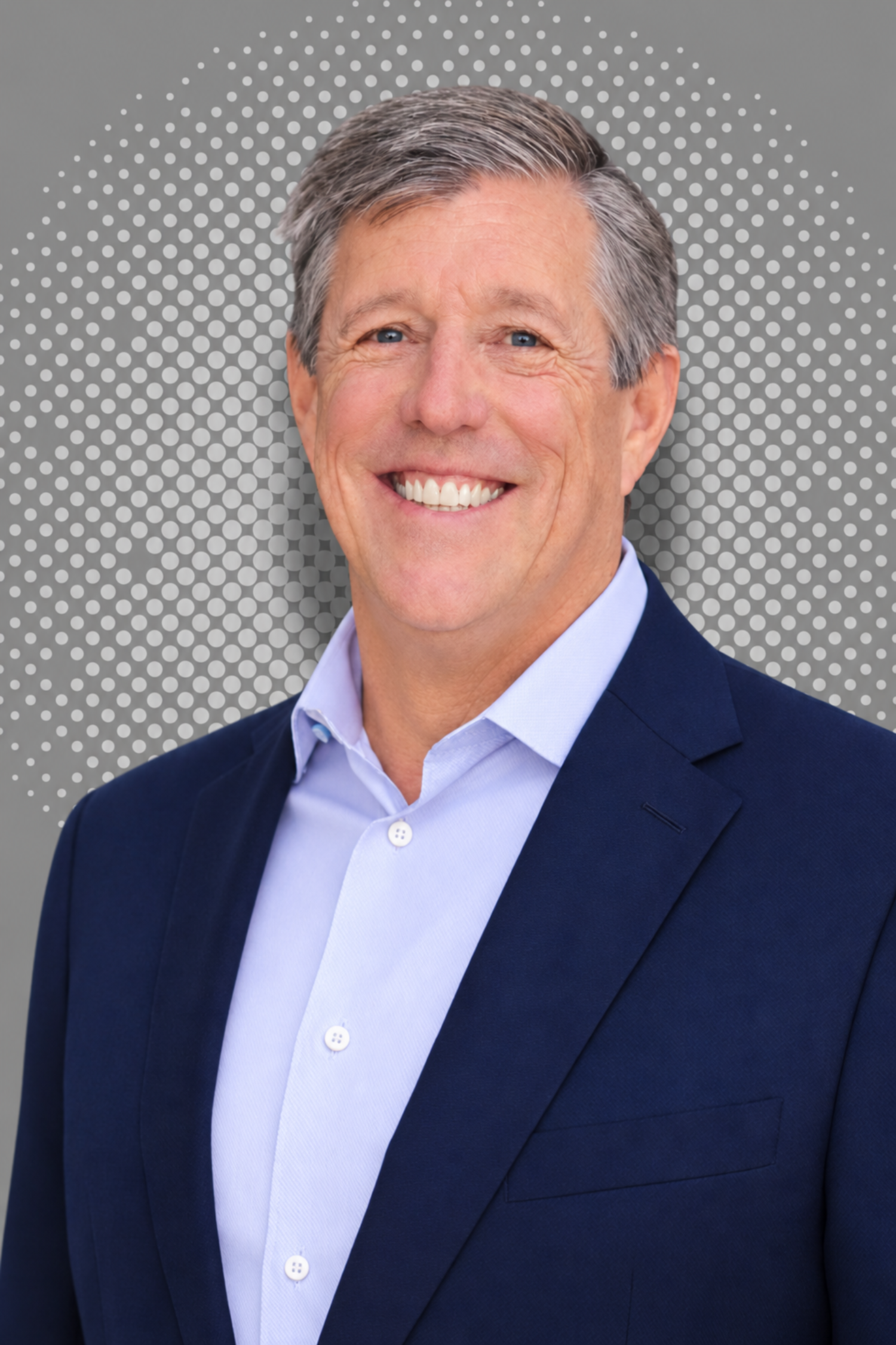 Smiling middle-aged man wearing a navy blazer and light purple dress shirt, standing against a gray background with a pattern of white dots.