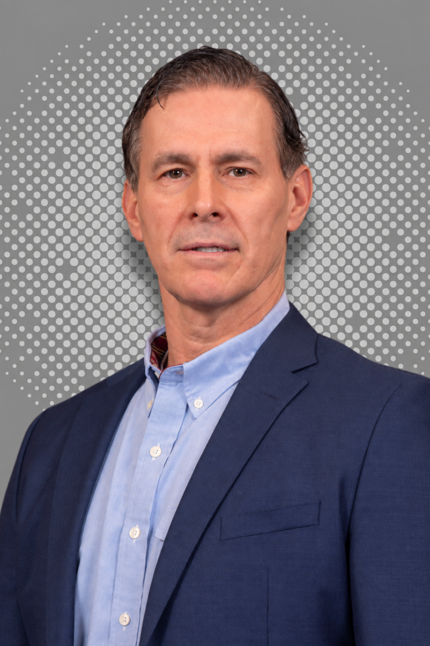 A middle-aged man in a blue suit and light blue shirt against a gray dotted background.