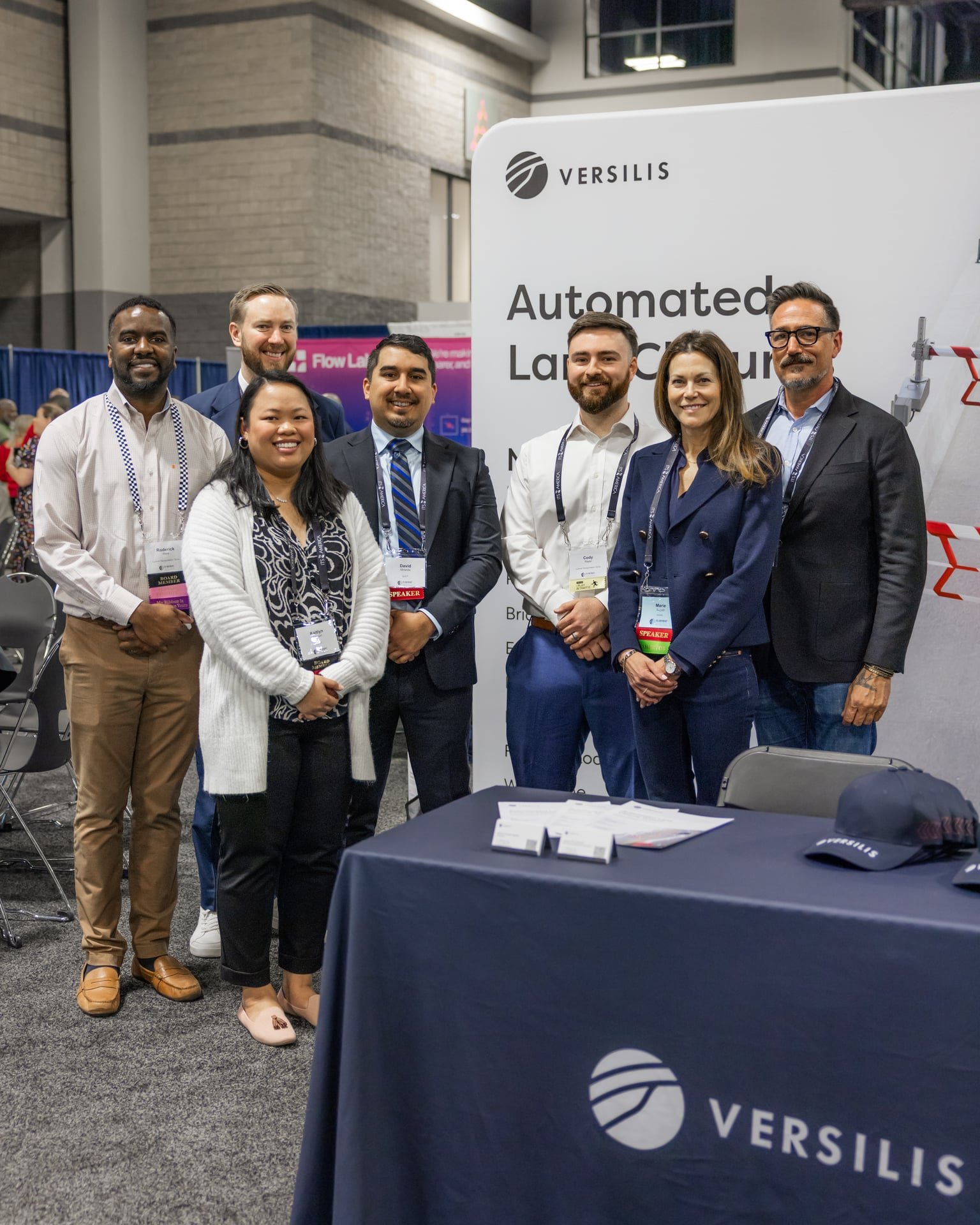 Group of seven diverse professionals standing behind a table at a conference, smiling for the photo. The table has a dark blue tablecloth with the Versilis logo, and a large white sign with Versilis branding and text that reads 'Automated Lab...' beh