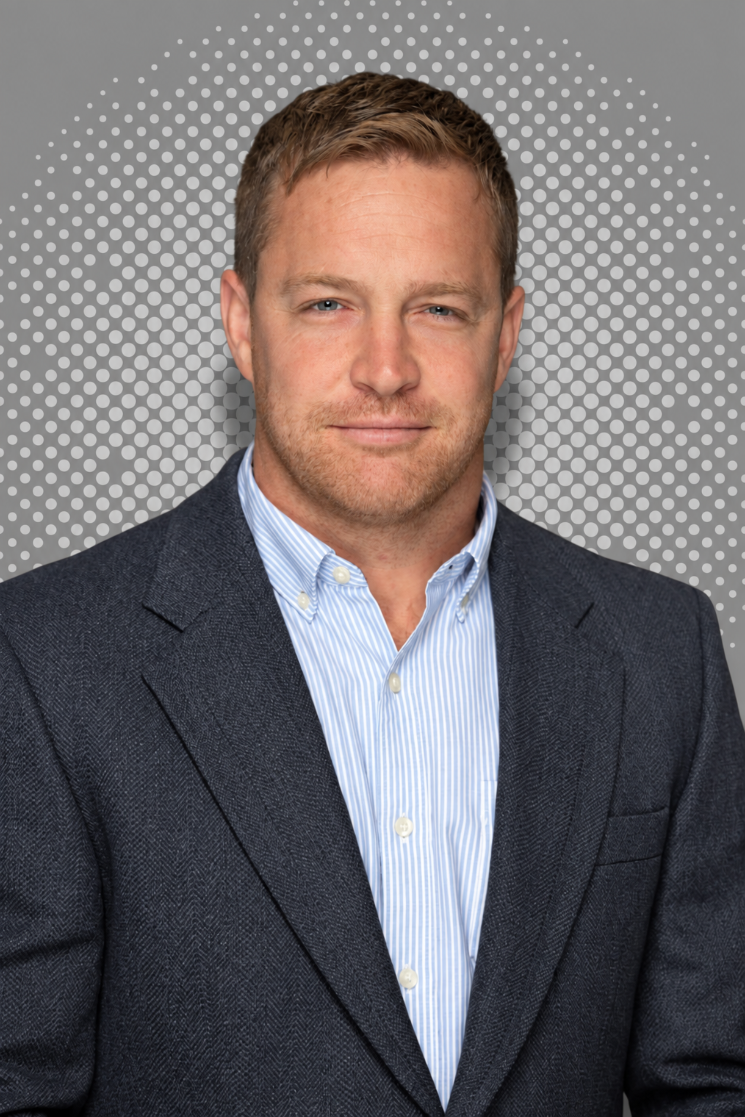 A man with light skin, brown hair, and blue eyes wearing a brown blazer over a striped shirt, smiling slightly against a background with white polka dots on a gray backdrop.