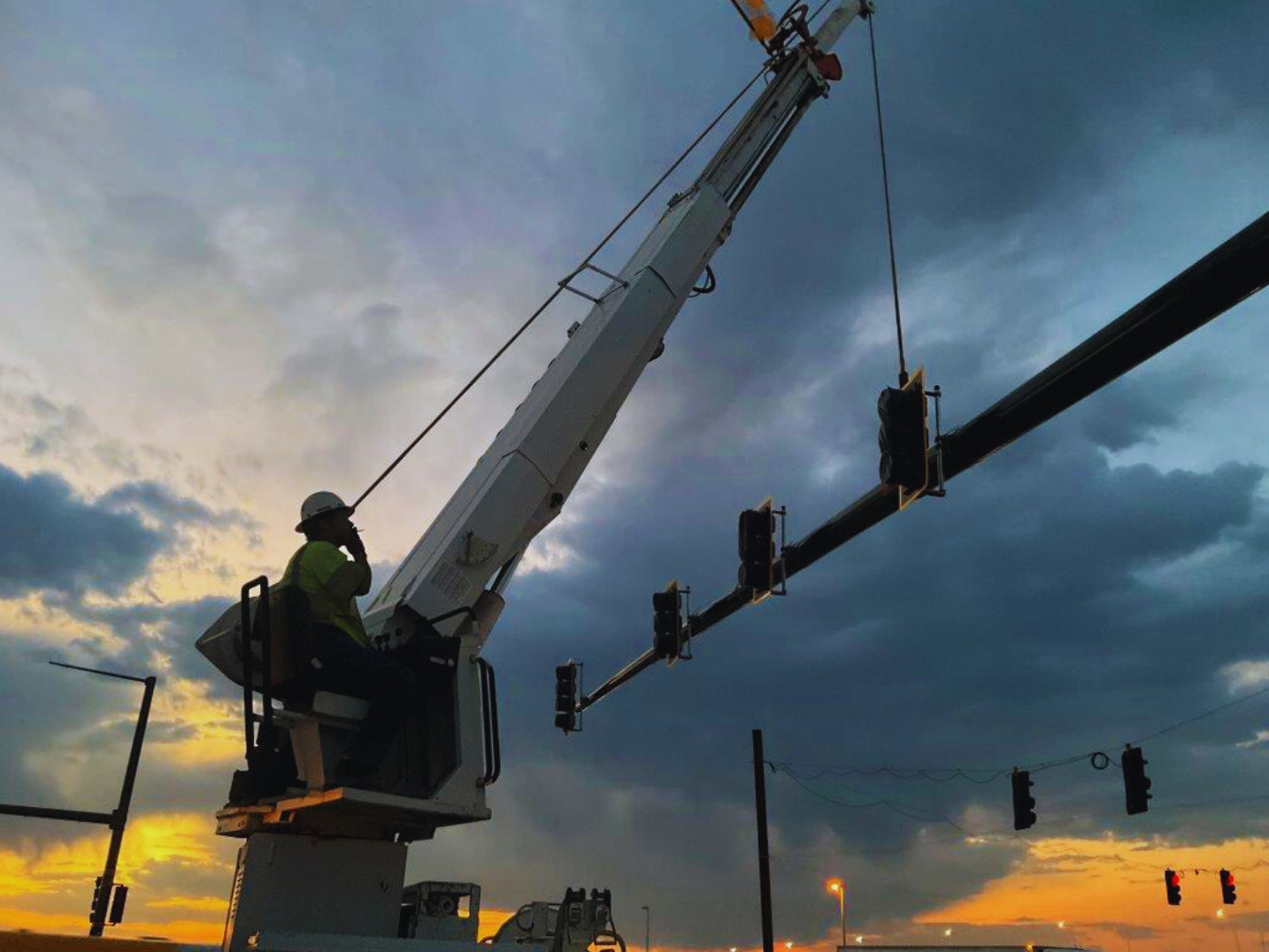 Worker in a cherry picker at sunset fixing traffic lights with a crane, under a cloudy sky.
