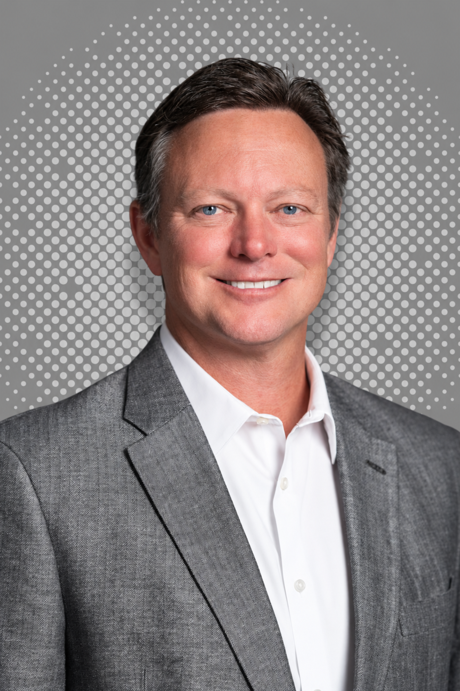 A headshot of a middle-aged man with short dark hair, blue eyes, wearing a grey suit jacket and white shirt, smiling, against a grey background with white dots.
