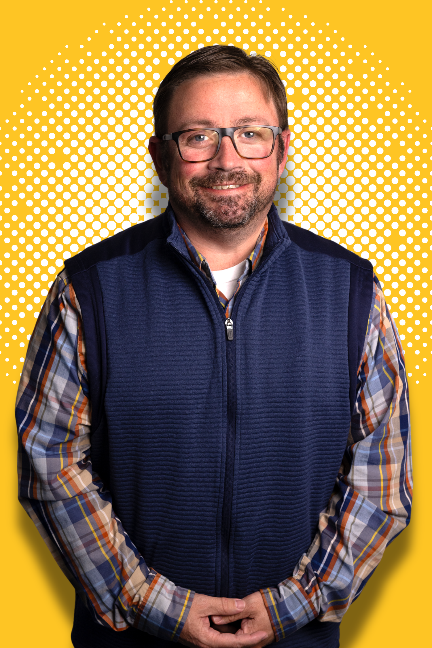 Man wearing glasses, plaid shirt, and blue vest against a yellow background with white dots.