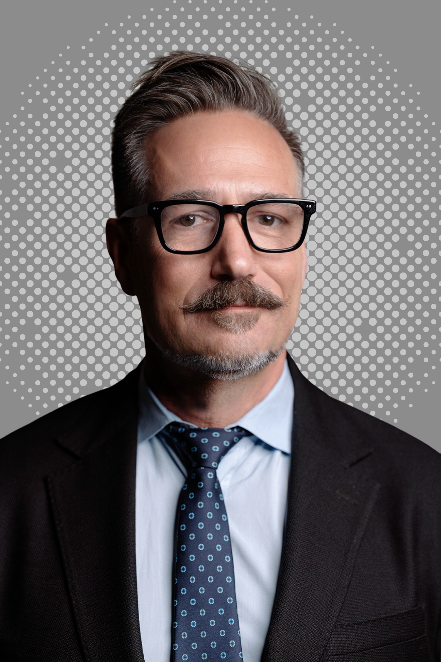 Man wearing glasses, navy suit, and white shirt against a gray dotted background.