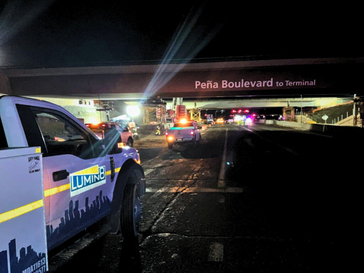 Nighttime scene under a bridge on Peña Boulevard, with work trucks and bright lights.
