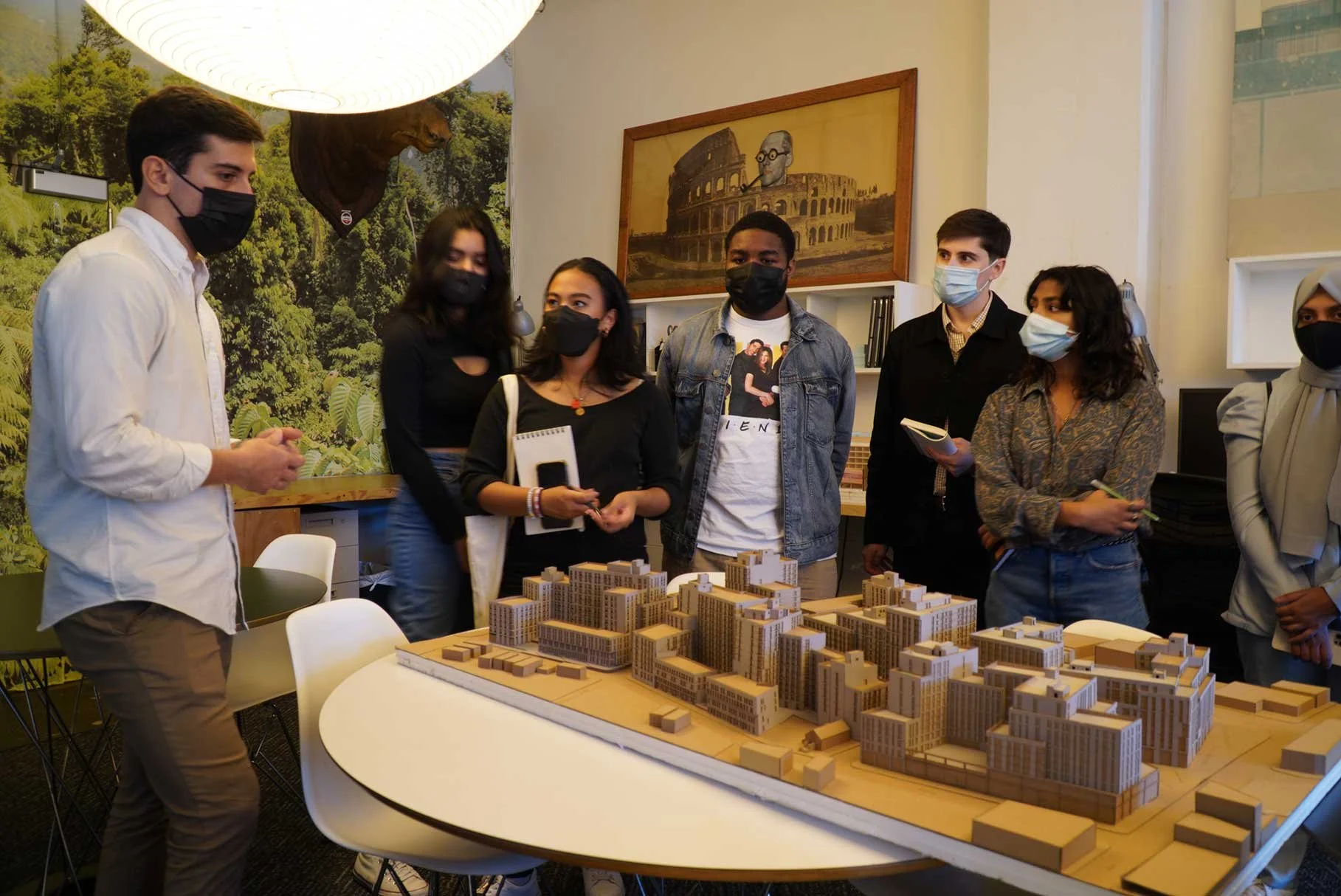 Group of young adults in a meeting room gathered around a detailed architectural model of a city block with buildings, discussing and taking notes, all wearing face masks.