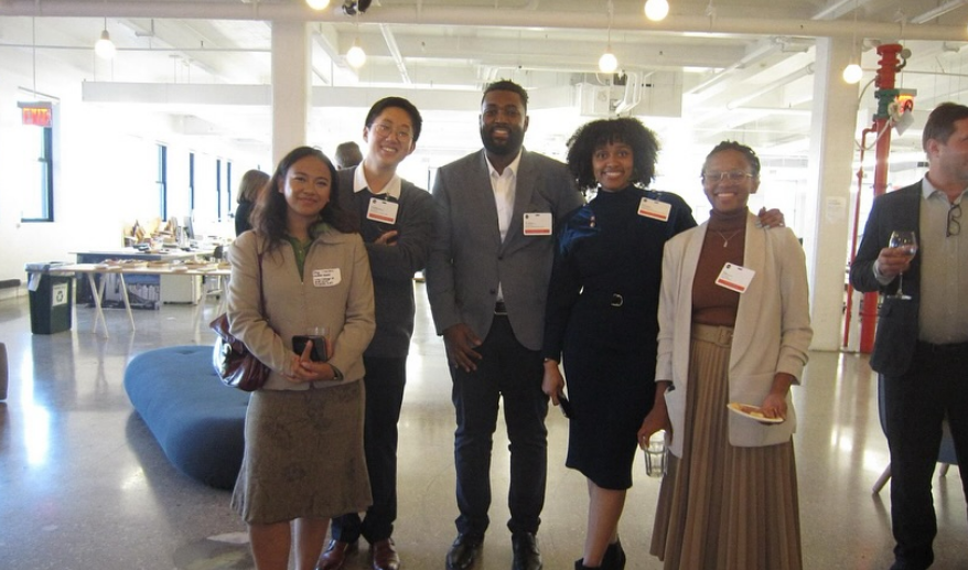 Group of five diverse professionals smiling and posing together at a social event in a modern office space, with some holding drinks or plates of food.