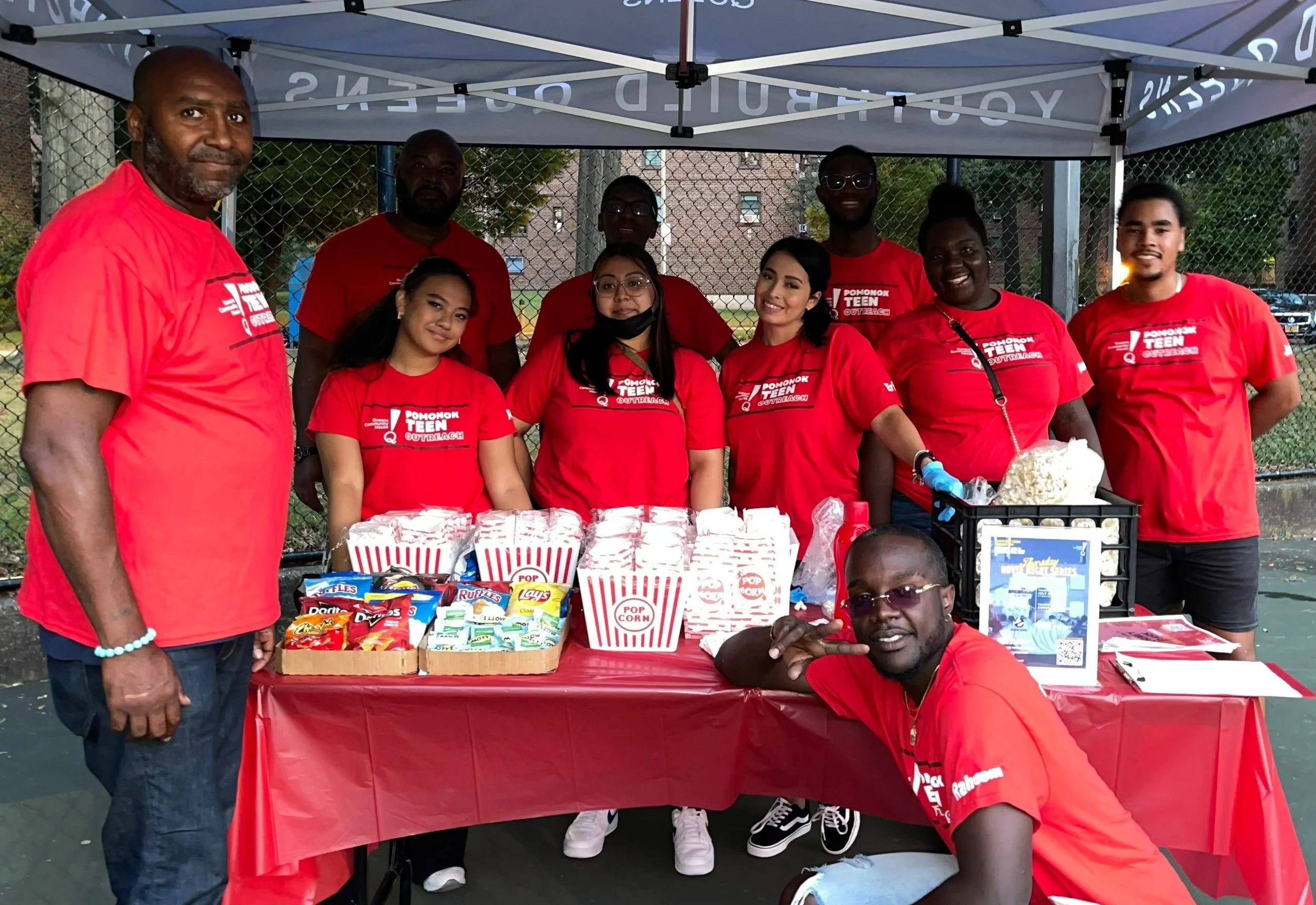 A group of eleven people wearing red shirts gathered around a table with popcorn and snacks at an outdoor event.