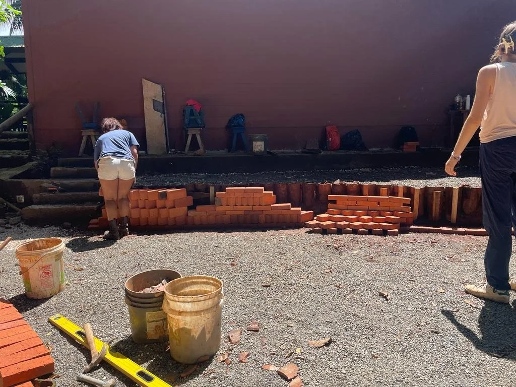 Two women are working on a bricklaying project outdoors. One woman, on the left, is leaning over and arranging bricks, while the other woman, on the right, stands nearby observing. There are construction tools, buckets, and stacks of bricks on the gr