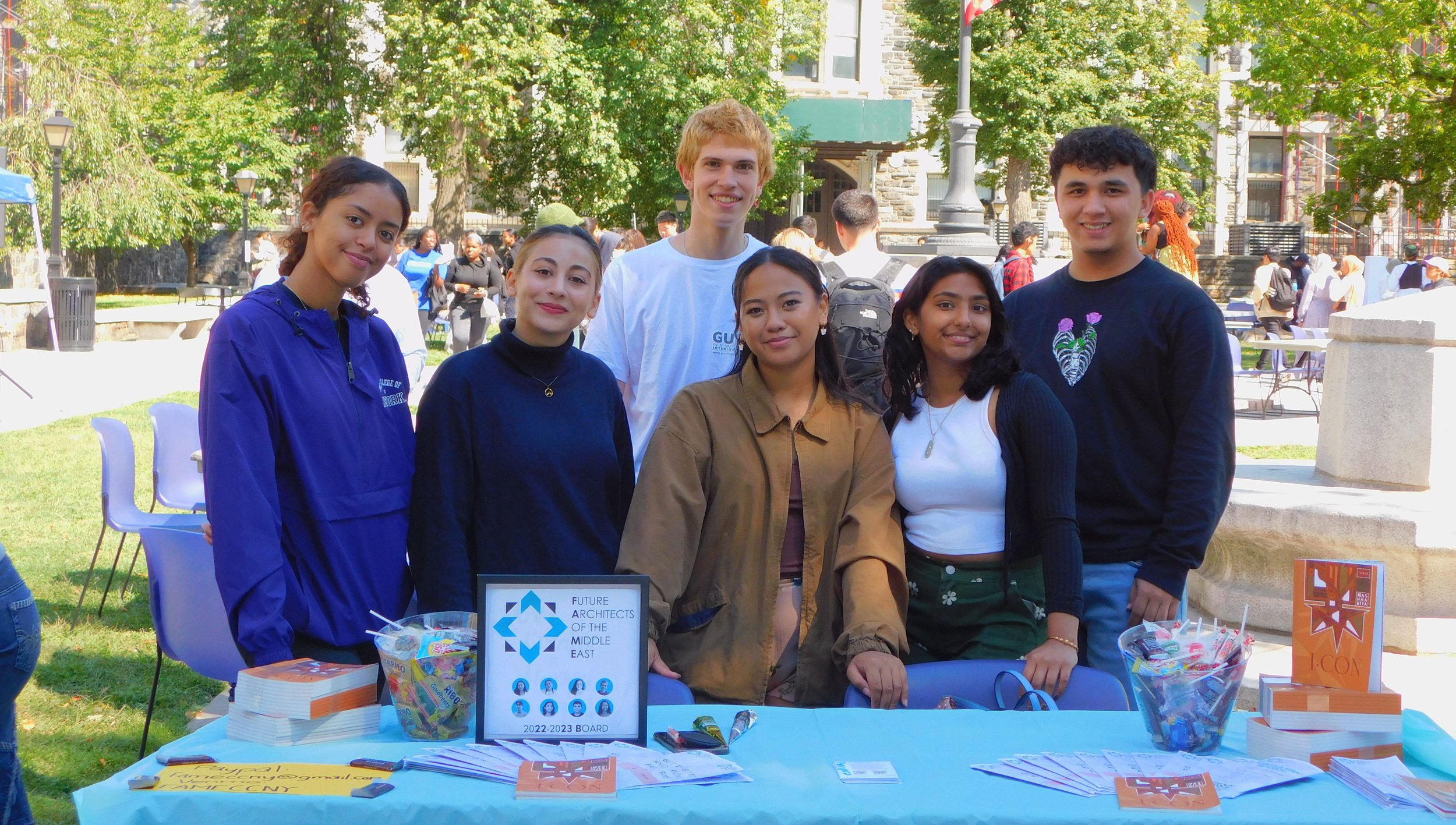 Group of six young people standing behind a table at an outdoor event, with trees, a stone building, and other attendees in the background.