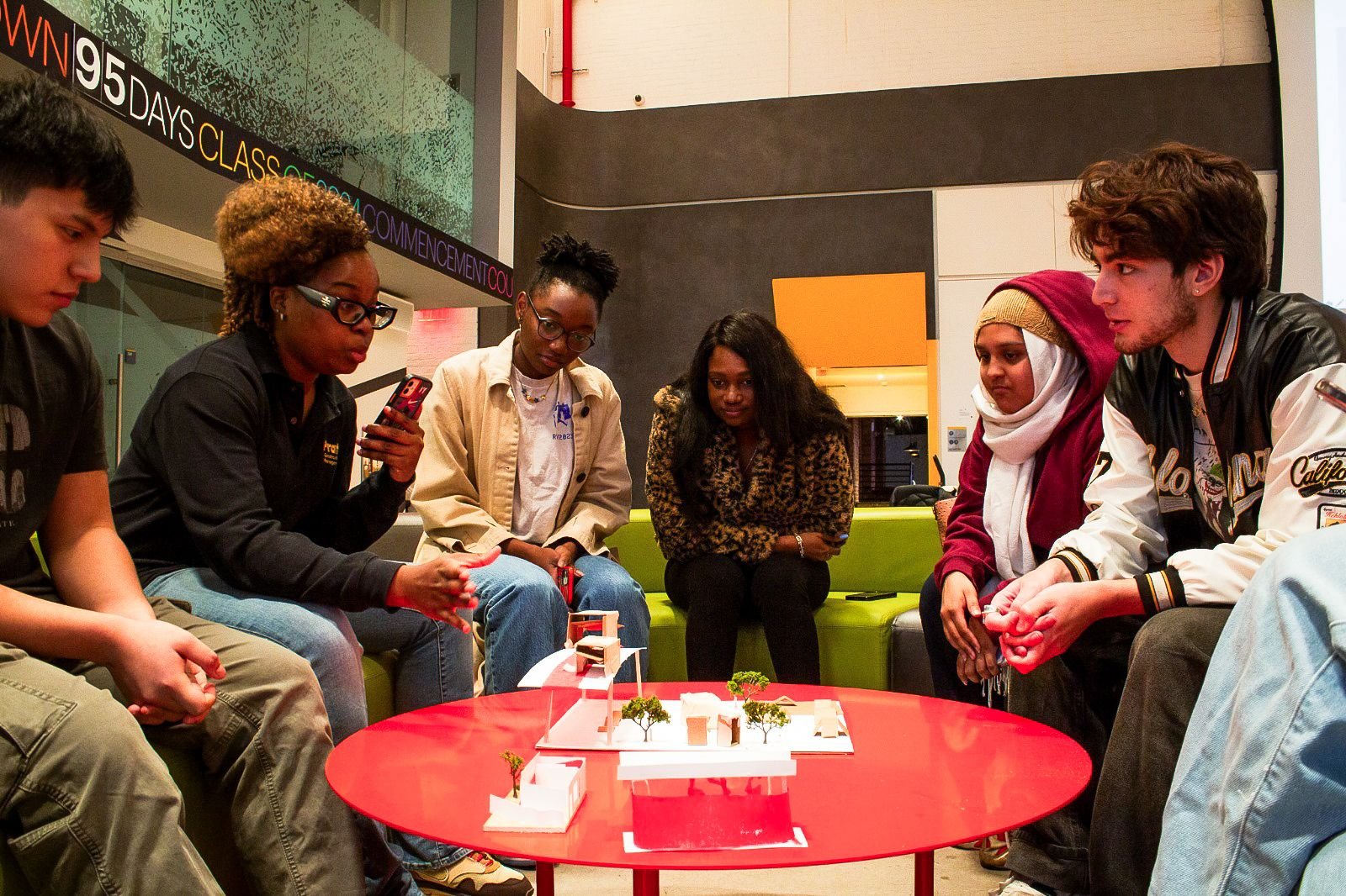 Group of diverse young people sitting around a red table, examining a scaled architectural model with trees, in a modern indoor space.