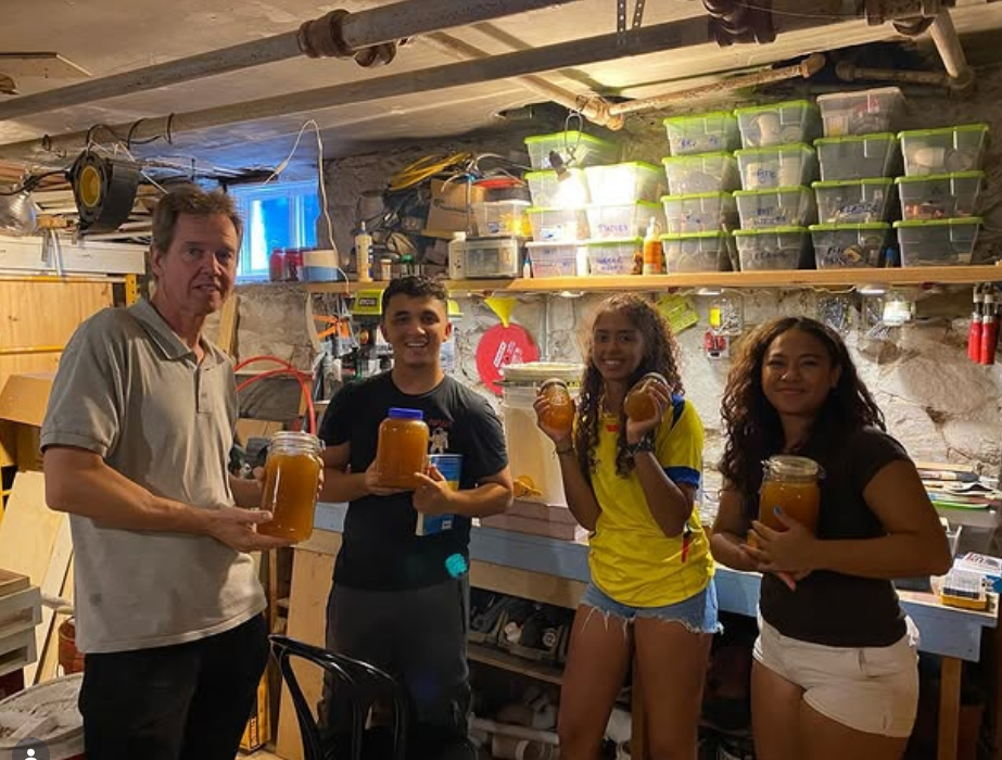 Four people standing in a cluttered basement workshop holding jars of orange substance, smiling at the camera.