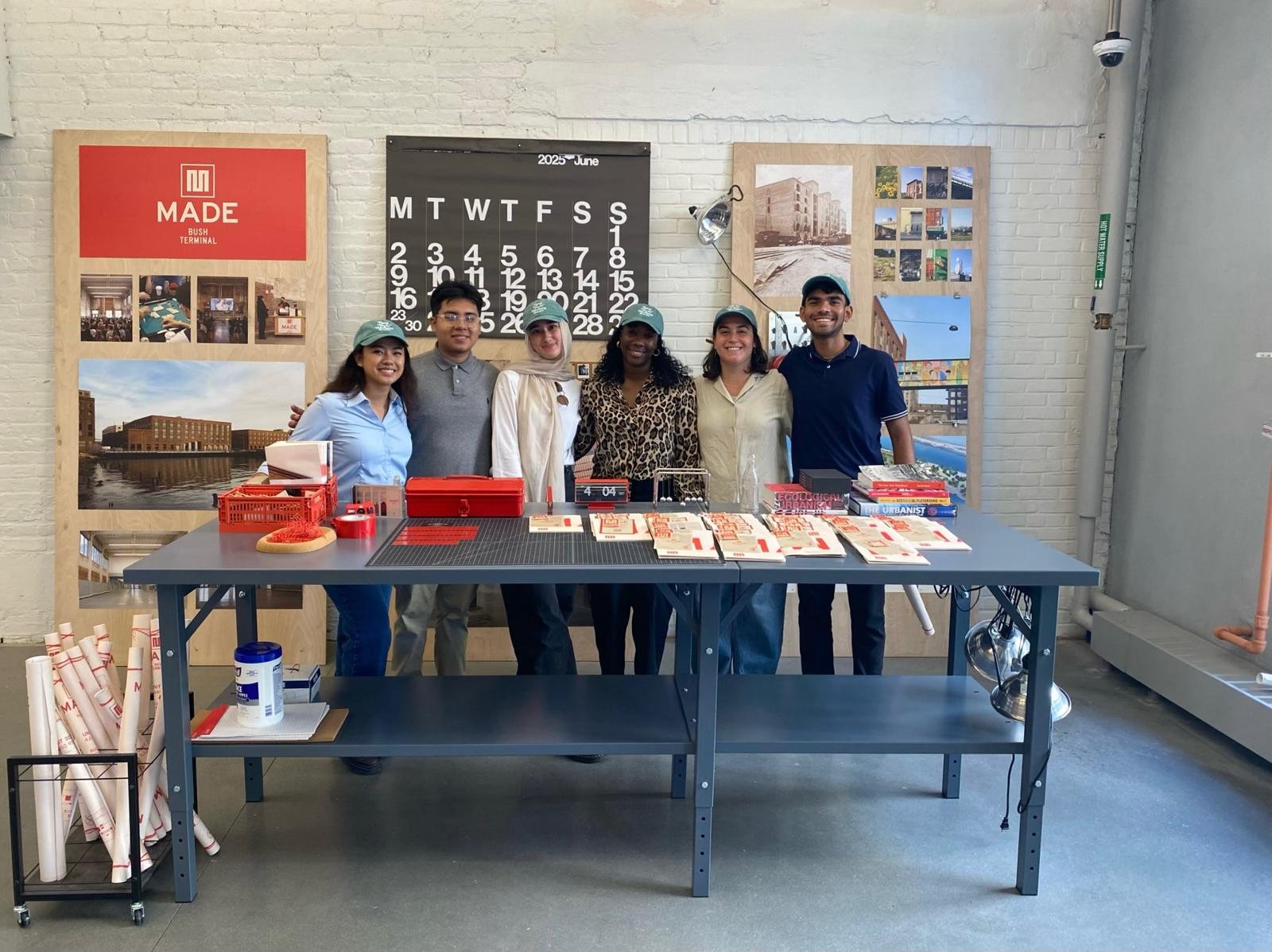 Six people standing behind a table filled with brochures and promotional materials inside a modern office space with white brick walls and large posters.