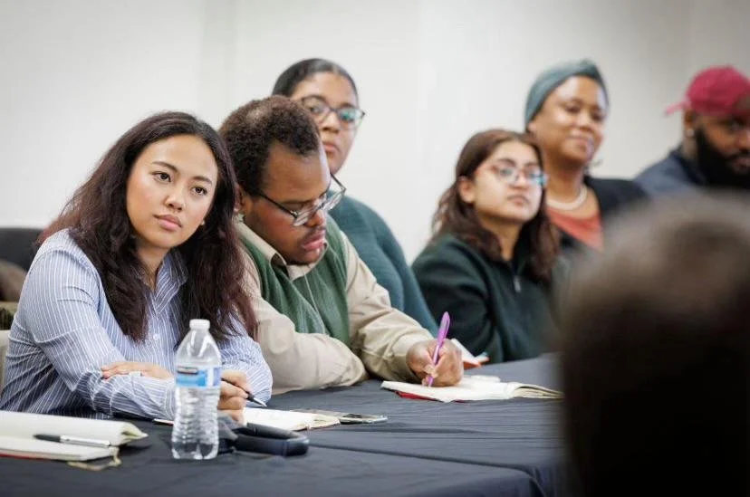 A diverse group of six adults sitting at a conference table, paying attention during a meeting or seminar.