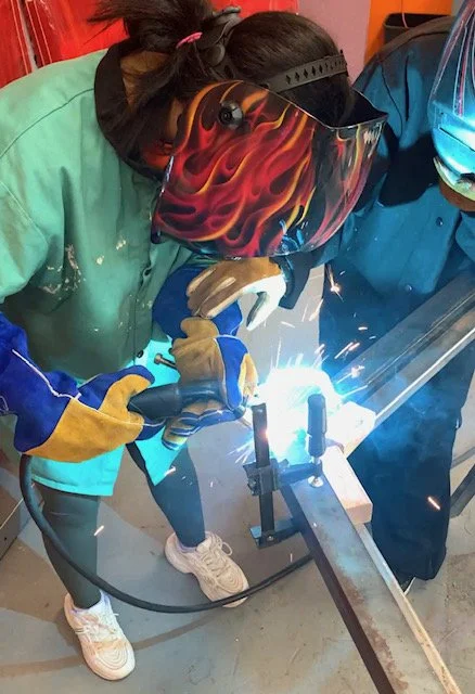 Two people welding metal pipes together in a workshop. One person is wearing a green shirt and yellow gloves, the other in a blue shirt, both with protective gear including a welding helmet and gloves.