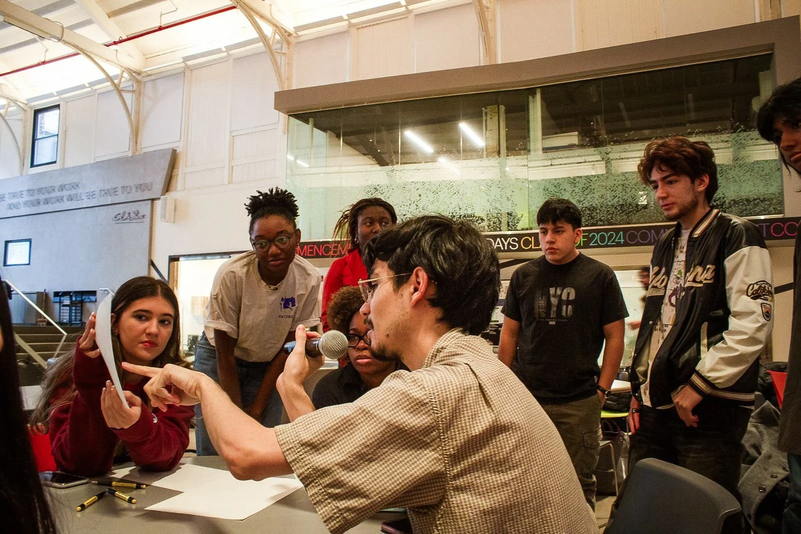 Group of teenagers gathered around a man with glasses holding a microphone, during a discussion or interview in a school or community center.