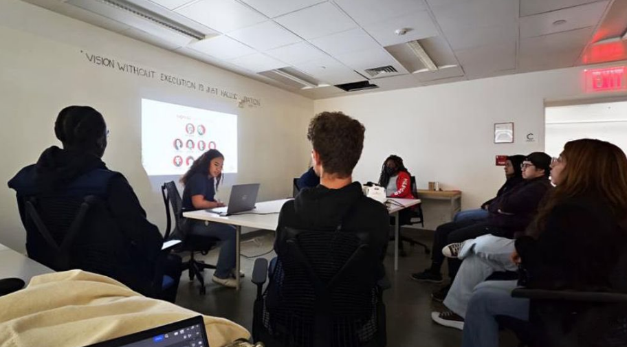 A group of people seated in a modern conference room, attending a presentation. The presenter, a young woman, is sitting at a table with a laptop and is speaking to the audience. The room has a white wall with the words 'VISION WITHOUT EXECUTION IS H