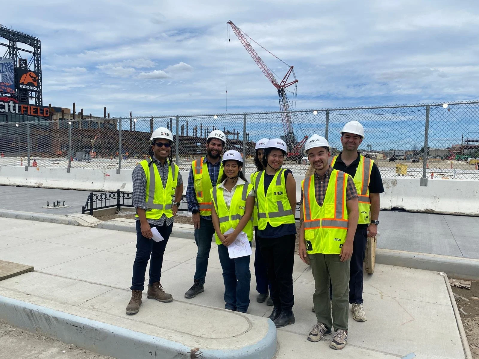 Group of construction workers wearing safety vests and hard hats standing on a sidewalk at a construction site with a crane in the background.