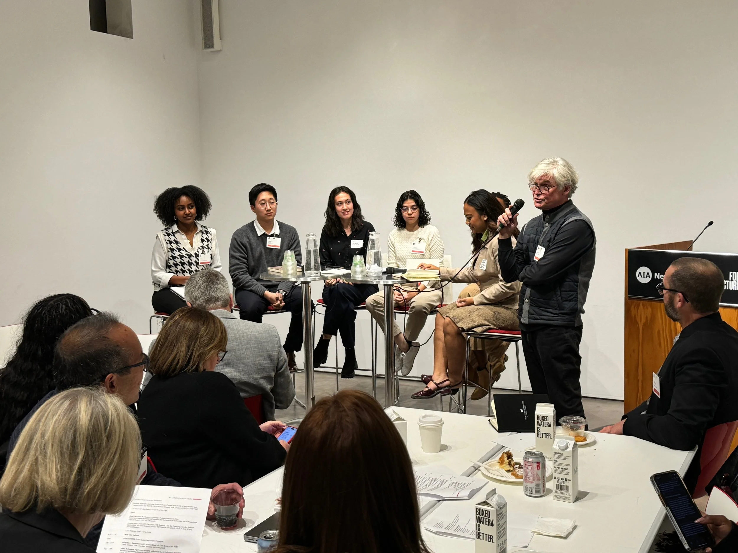 A panel discussion with five women seated and one man standing and speaking into a microphone at conference. Audience members are seated at tables with papers, drinks, and food.