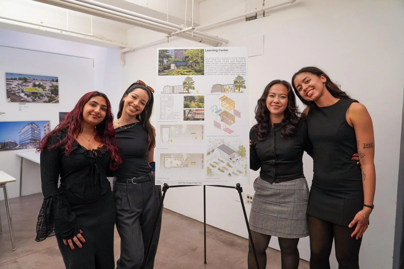Four women stand together and smile in front of a research poster in an indoor setting. The poster contains images, diagrams, and text related to a learning center project. The women are dressed in black and gray, and the room has white walls with ad