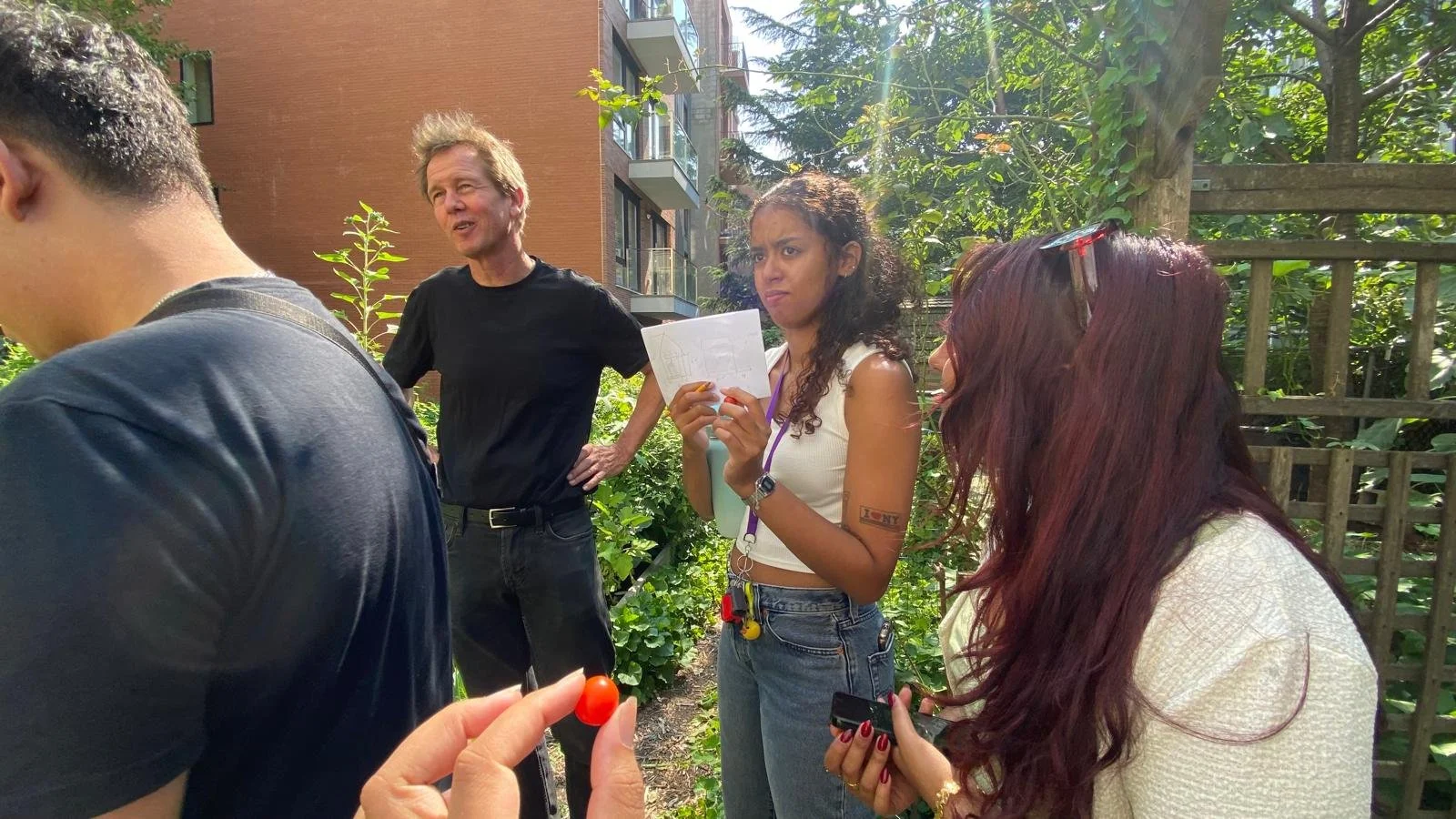 A group of four people outdoors in a garden, engaging in conversation. A man with blonde hair is talking, while a woman with curly hair and a tank top holds a notepad. Another woman with long red hair and glasses on her head is looking down at her ph