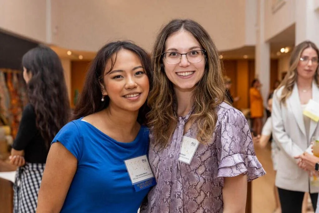 Two women smiling at a professional event, standing close together with name tags, in a brightly lit room with other people in the background.