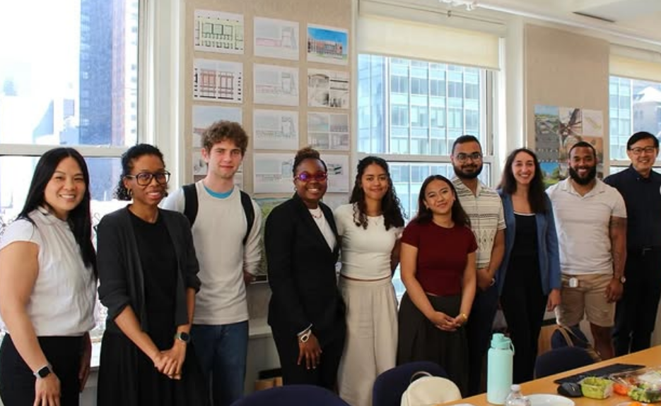 Group of ten diverse young adults standing inside a modern office with large windows and cityscape view, smiling for a photo.