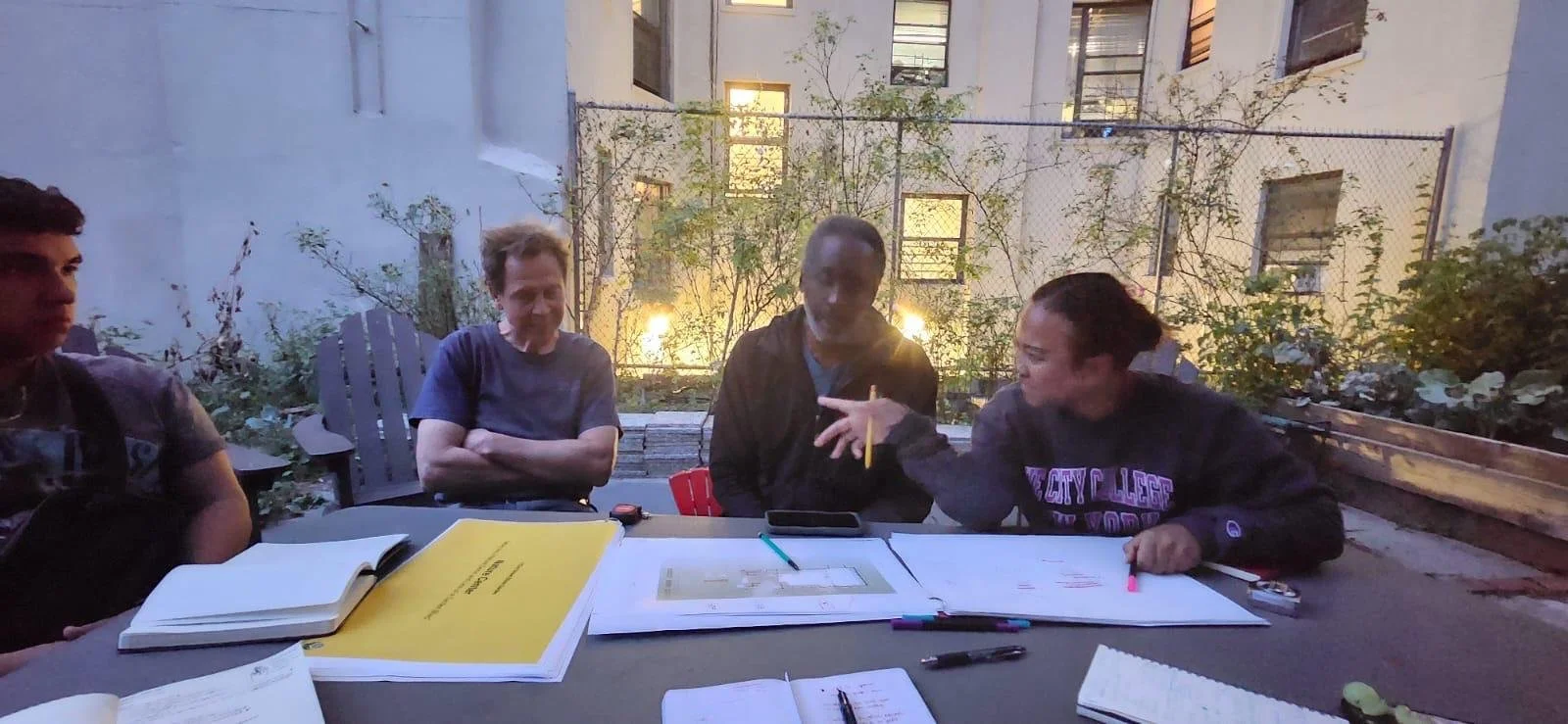Four people sitting at a table outside during the evening, engaging in a discussion with open notebooks, papers, and pens on the table. One woman is talking and gesturing with her hand, while others listen attentively, with a building and some greene