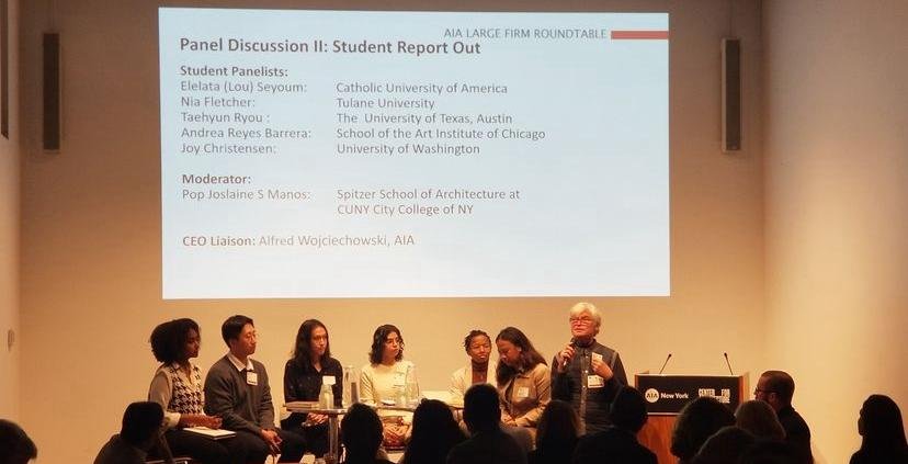 Panel discussion at a conference with six speakers seated side by side on stage, and a man standing and speaking. A large screen behind them displays the panel details, including names of student panelists from various universities, a moderator, and 