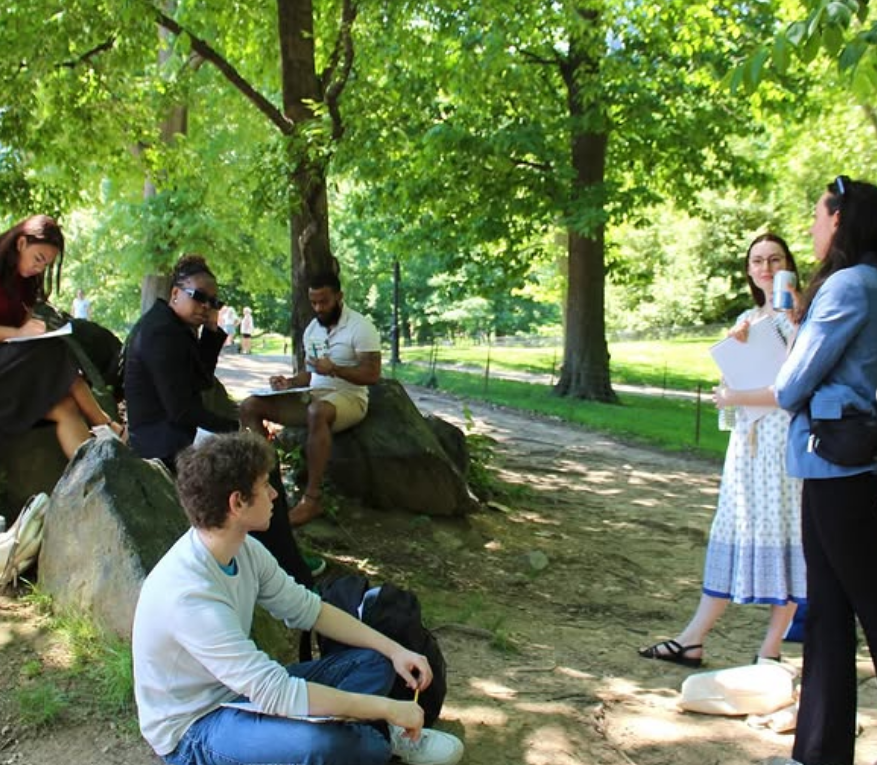 Group of young adults sitting and standing in a shaded park, some taking notes or holding notebooks, engaging in a conversation on a sunny day.