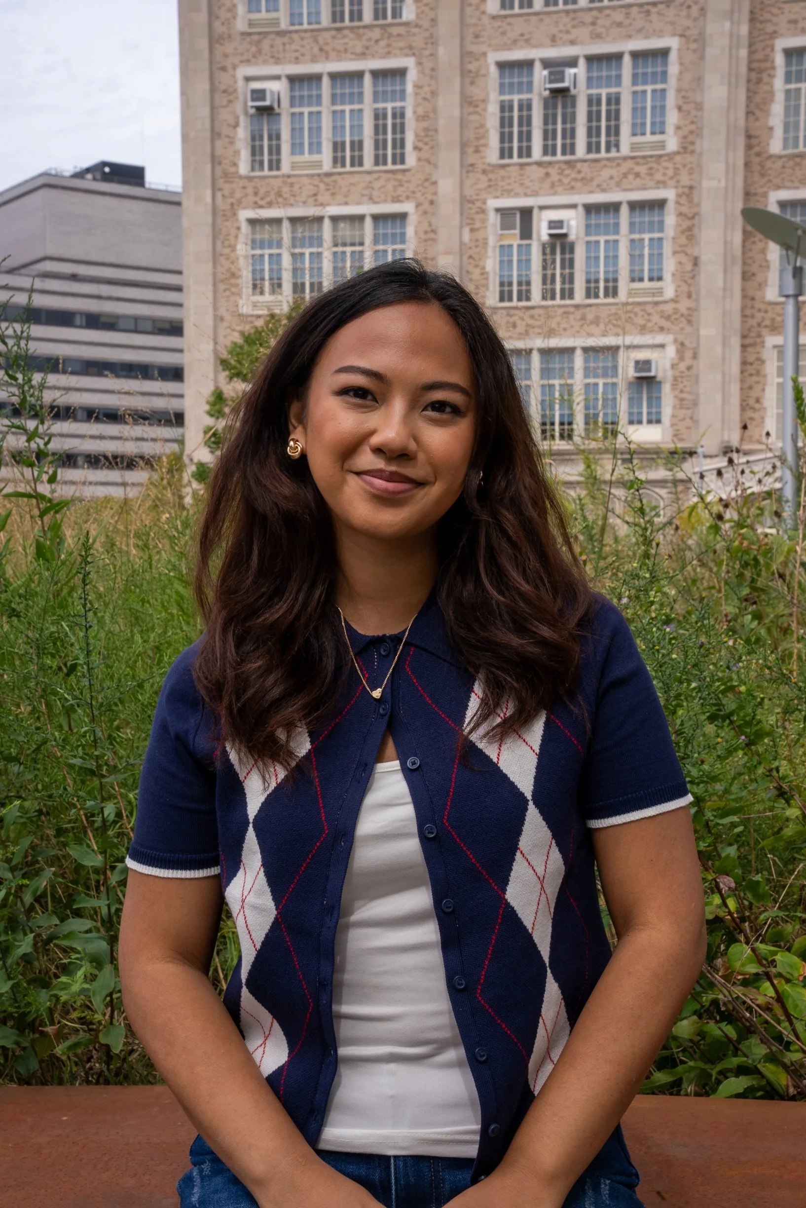 A woman with long dark hair wearing a navy blue and white argyle cardigan, standing outdoors with greenery and a building in the background.