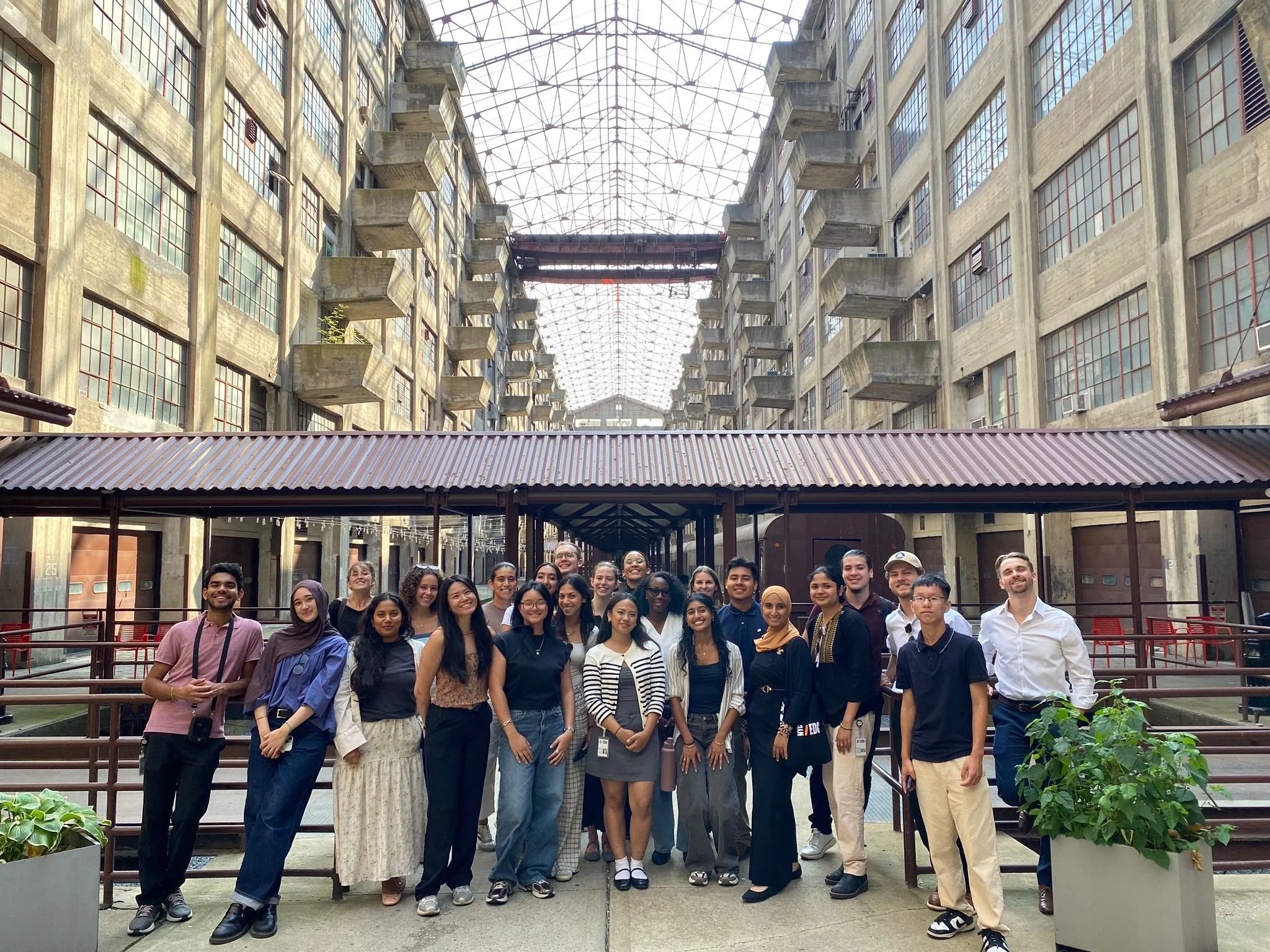 Group of diverse people posing together outdoors in front of a building with a glass ceiling and concrete walls, some with balconies, during the daytime.