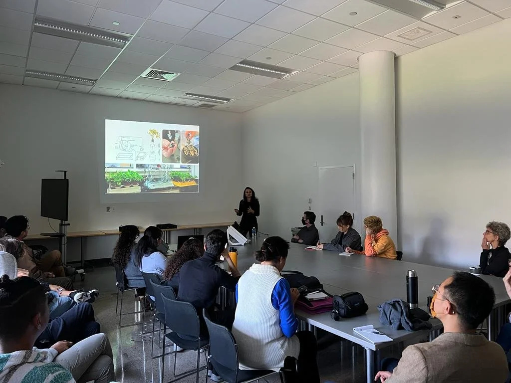 A woman is giving a presentation in a conference room to a group of people seated around a large table. The presentation is projected onto the white wall behind her, showing various images related to plants, food, and possibly science or technology.